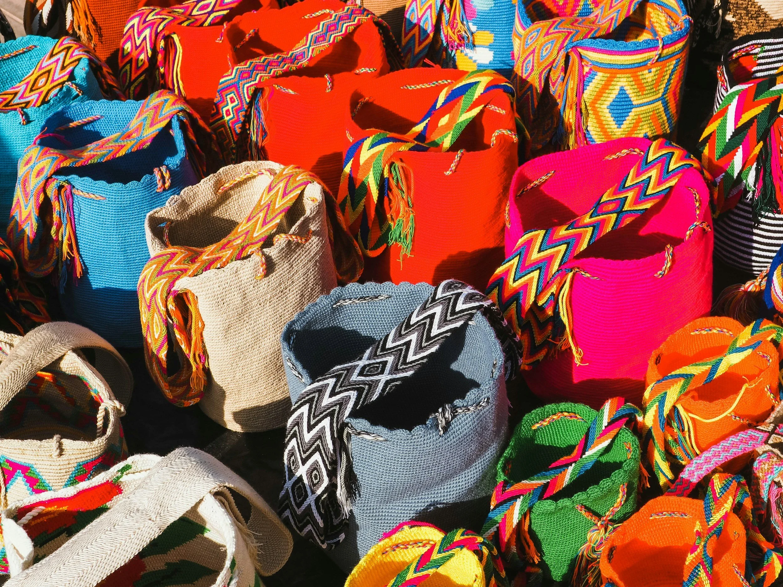 Colorful woven bags with intricate geometric patterns and bright colors such as red, pink, blue, green, orange, and beige displayed outdoors.