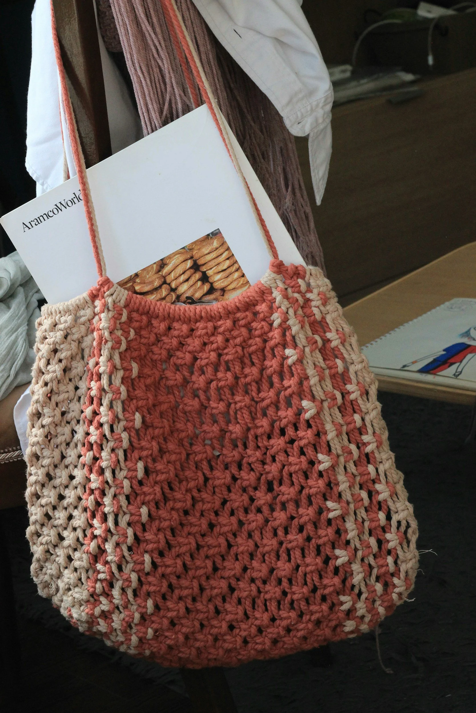 Pink and cream crochet bag hanging on a wooden chair, with documents and a magazine inside the bag. The magazine has a picture of cookies on the cover.