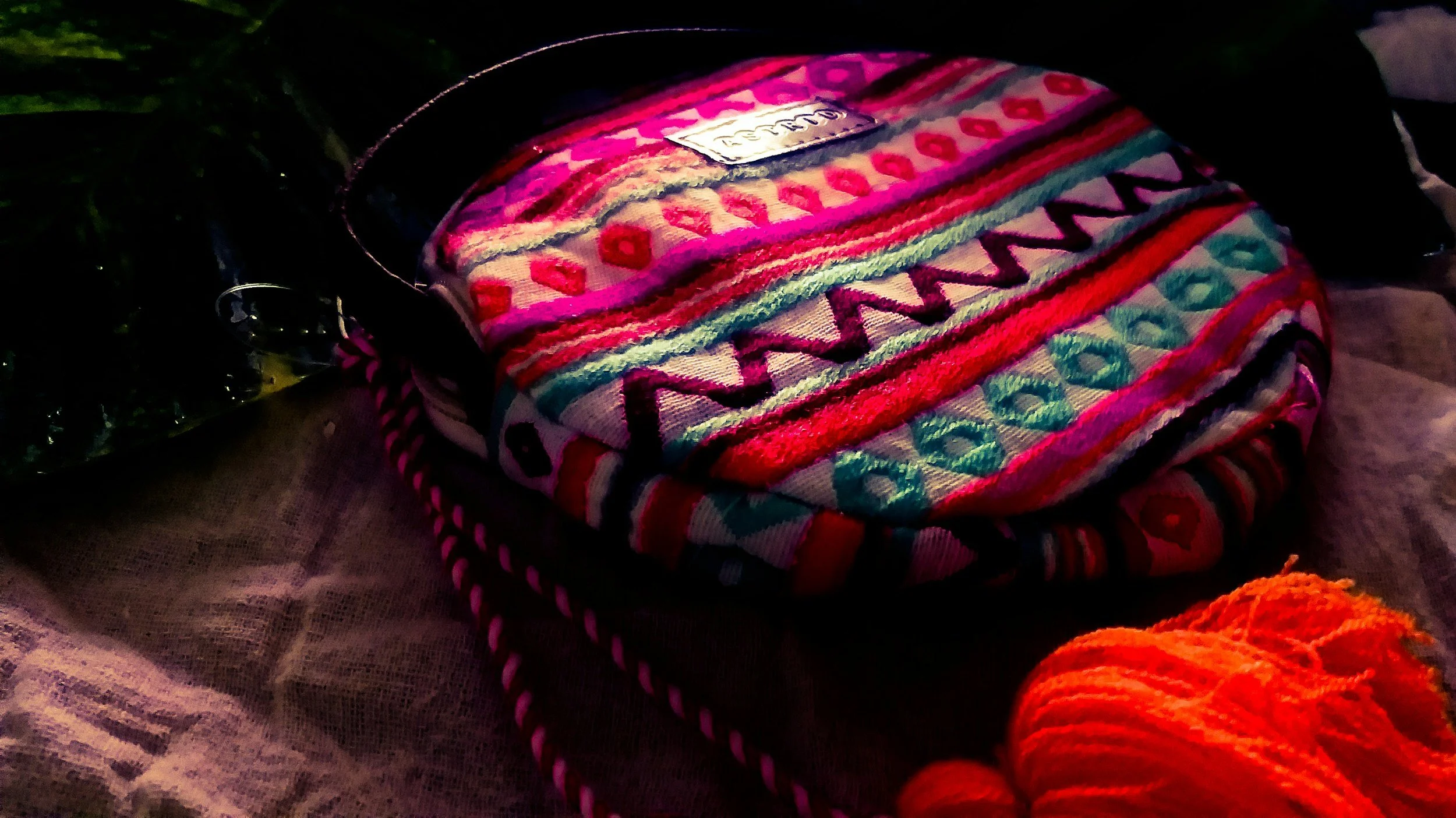 Colorful woven bag with geometric and heart patterns resting on a textured surface, with a hint of orange yarn or fabric in the foreground.
