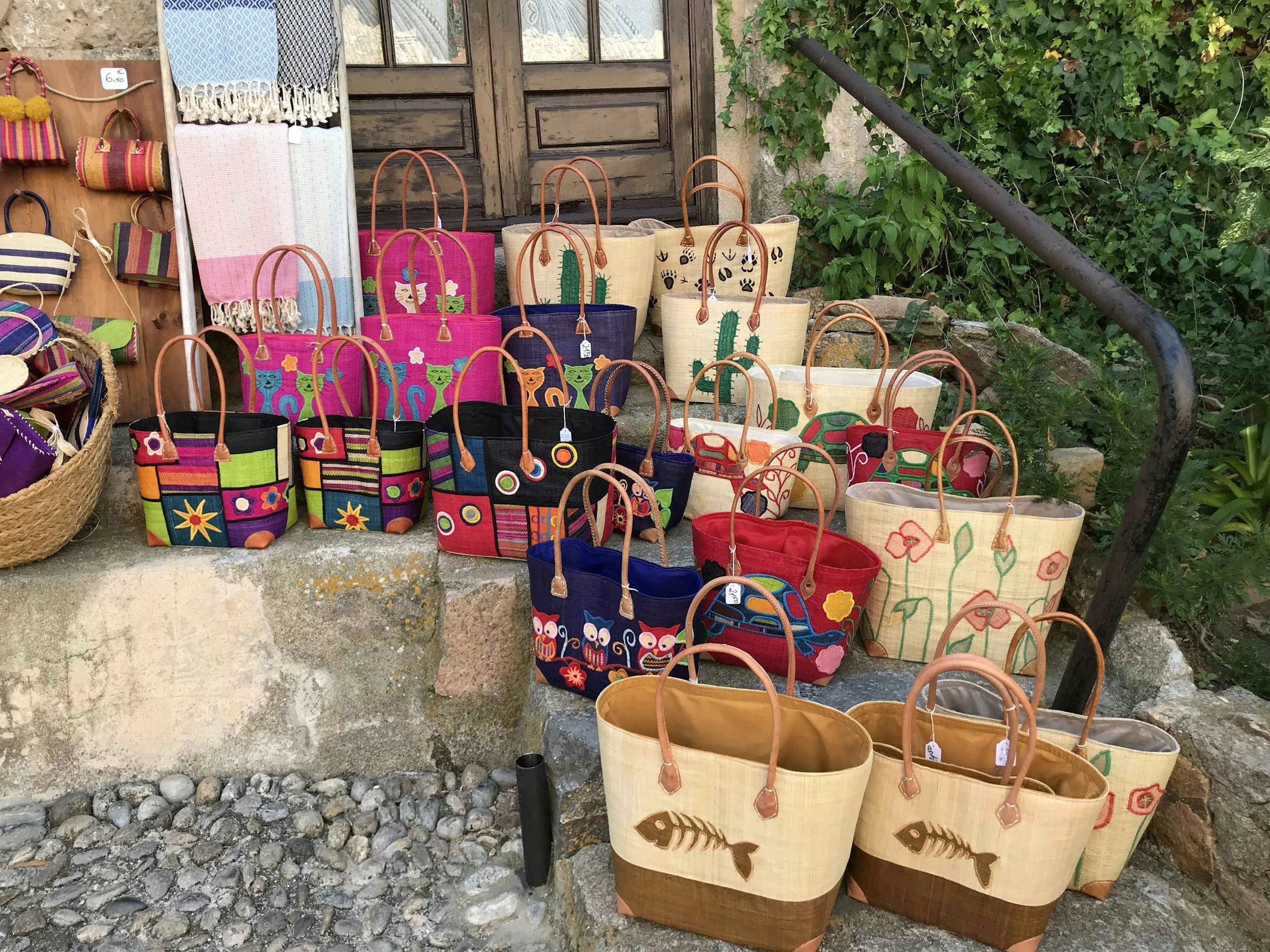 Colorful woven tote bags with animal and plant designs displayed outdoors near a stone wall and green bushes.