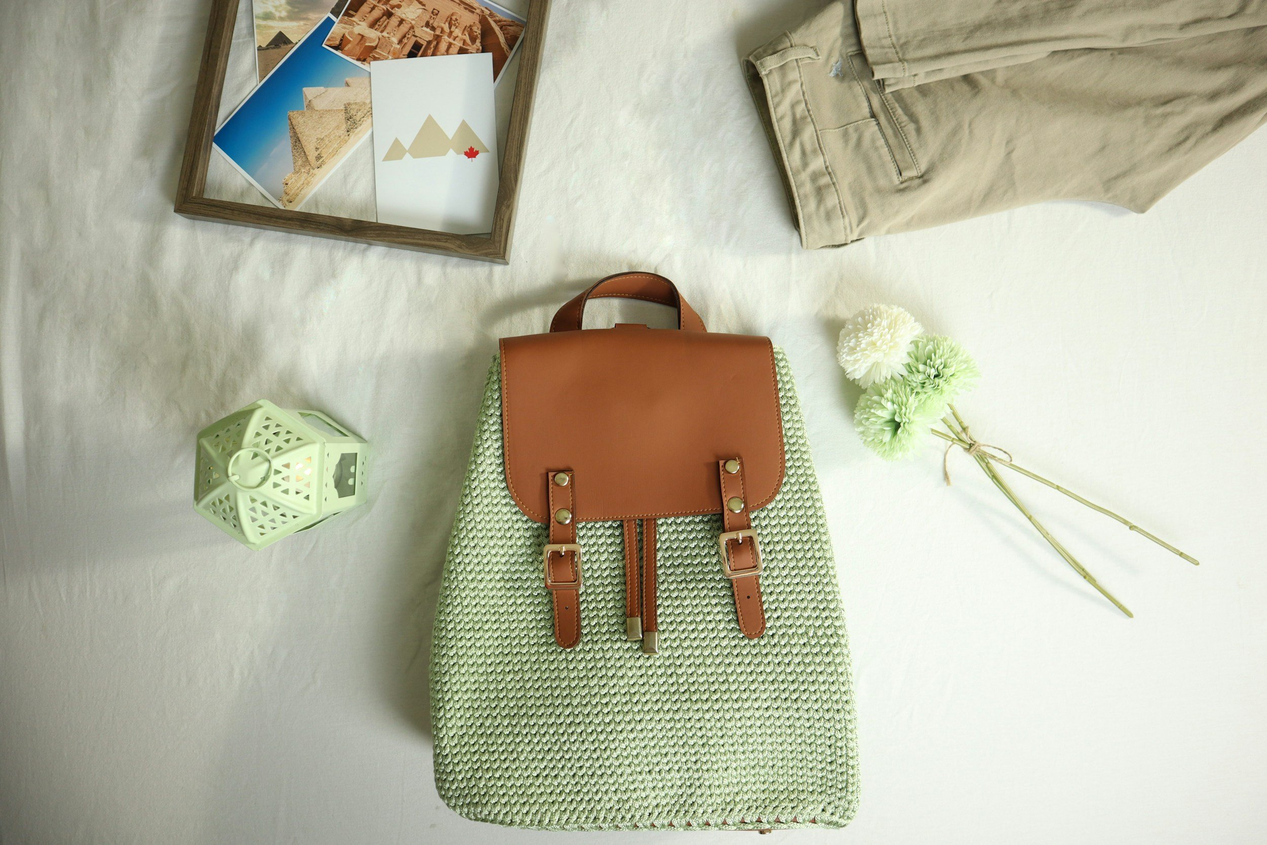 Flat lay of a collection of items on a white surface, including a framed postcard of Egyptian pyramids, a white postcard with a mountain logo and red maple leaf, khaki pants, a brown leather handbag, a green textured scarf, a small green decorative lantern, and a bouquet of white and light green flowers.
