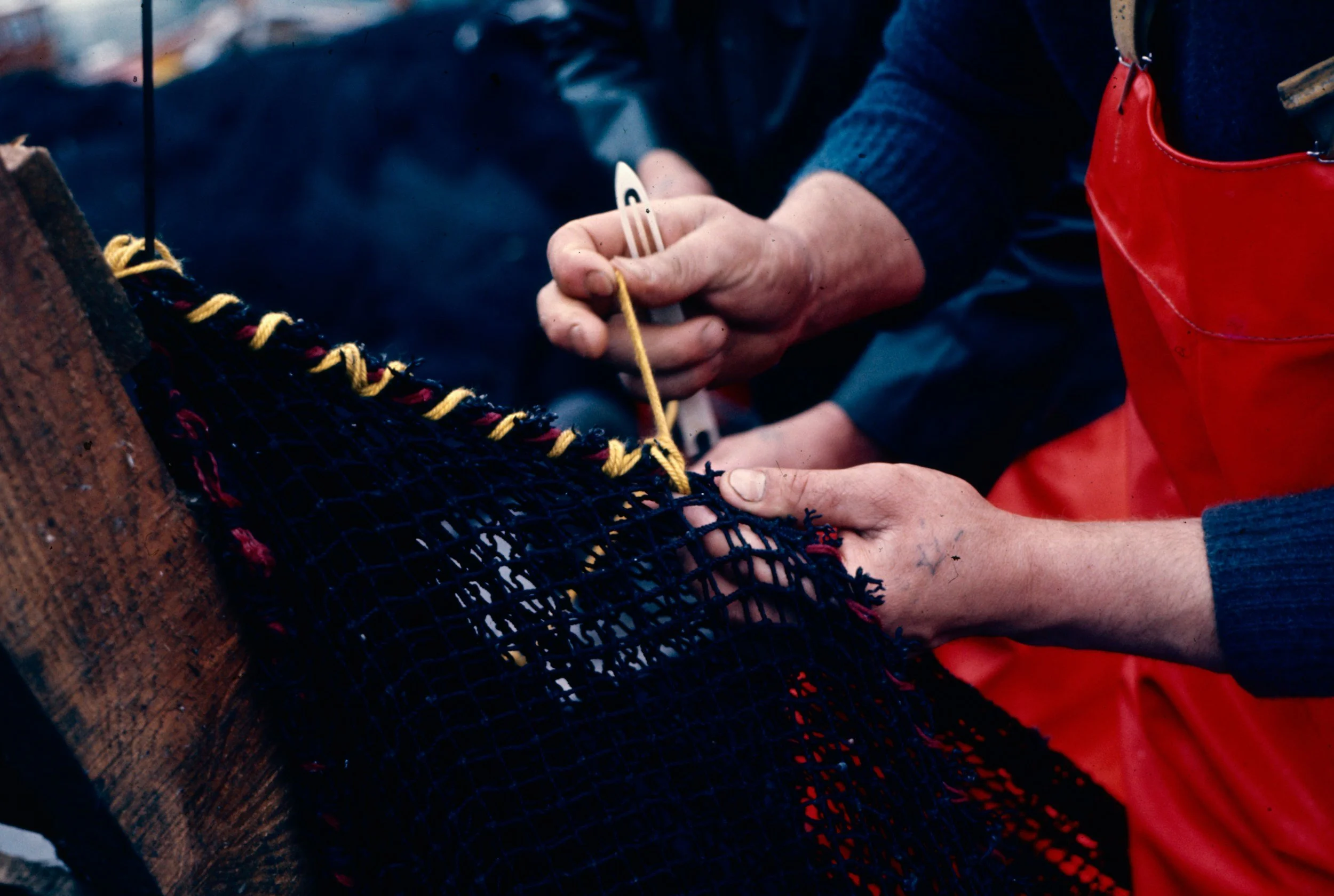 Close-up of a person's hand sewing or knitting dark-colored netted fabric with yellow and red threads, while another person, wearing a red apron, holds the fabric.