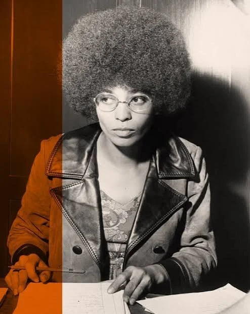 A woman with a large afro hairstyle, wearing glasses and a leather jacket, sitting at a table with papers in front of her, in a black and white photo with a colored vertical strip on the left side.