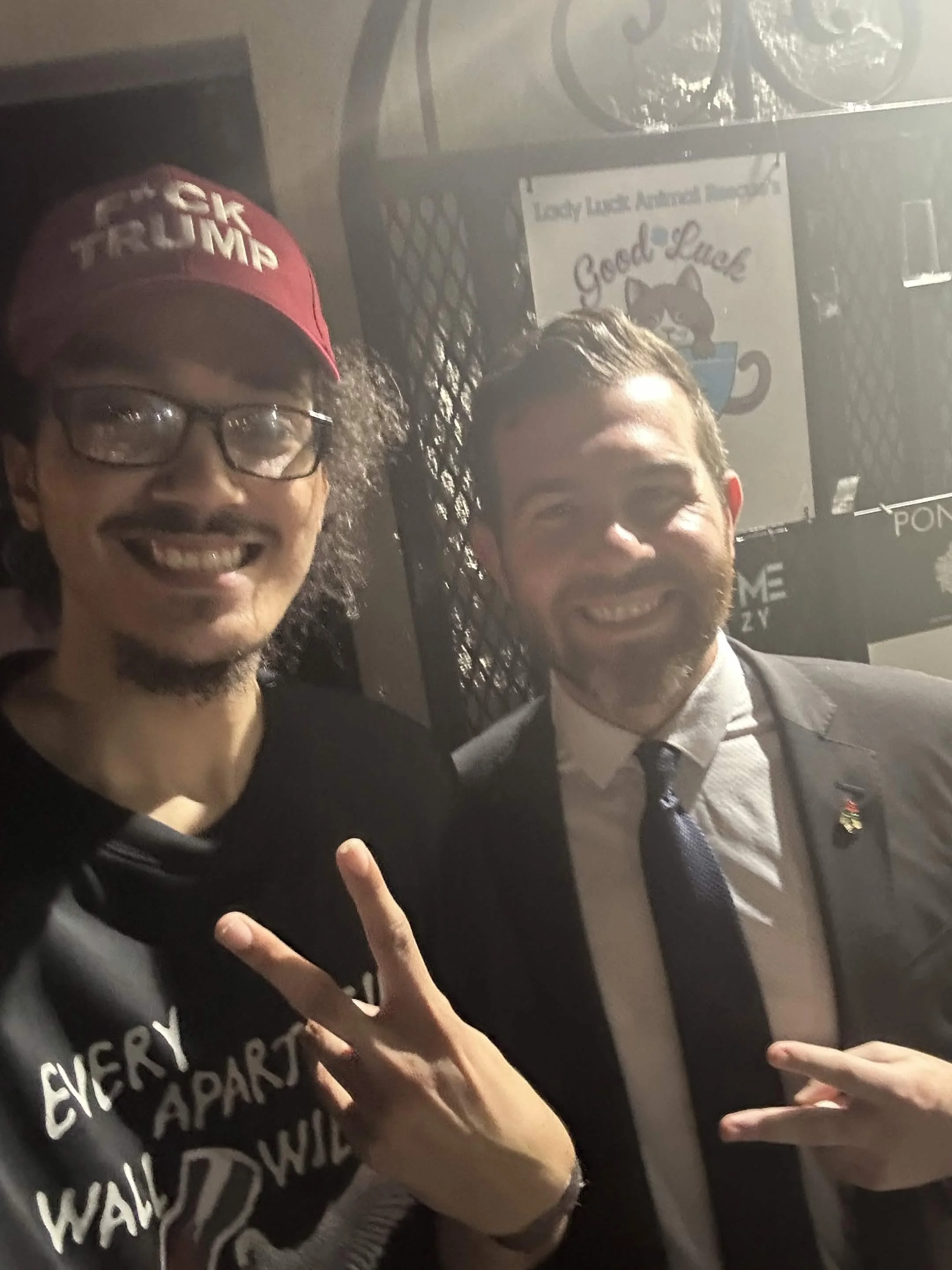 Two men smiling and posing for a selfie; one wears a red 'Make America Great Again' hat and glasses, the other in a suit and tie, with a hand showing a peace sign.