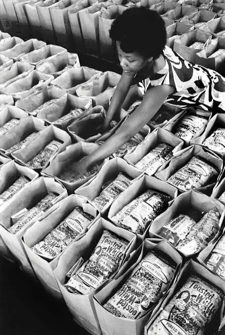 A young woman with short, curly hair reaches into a box among many open boxes filled with bread loaves at a bakery or grocery store.