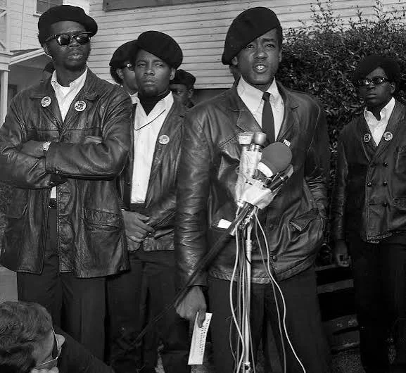 A group of African American people, including women and men, standing outdoors in front of a wooden building. They are wearing matching leather jackets and berets. One woman is speaking into a microphone, possibly giving a speech or statement, while others stand behind her, some with arms crossed and serious expressions.