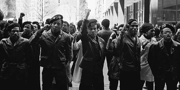 A black-and-white photo of a group of women protesting on the street, some raising their fists. They appear determined and united in their cause.