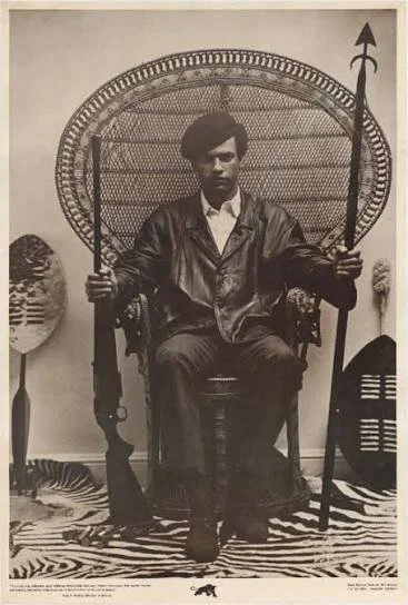 A young man in a leather jacket sitting on an ornate chair with a high back, holding a spear and a rifle, posed in a studio with animal skins and carved objects as decoration.