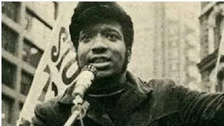 Black and white photo of a person speaking into a microphone on a city street.