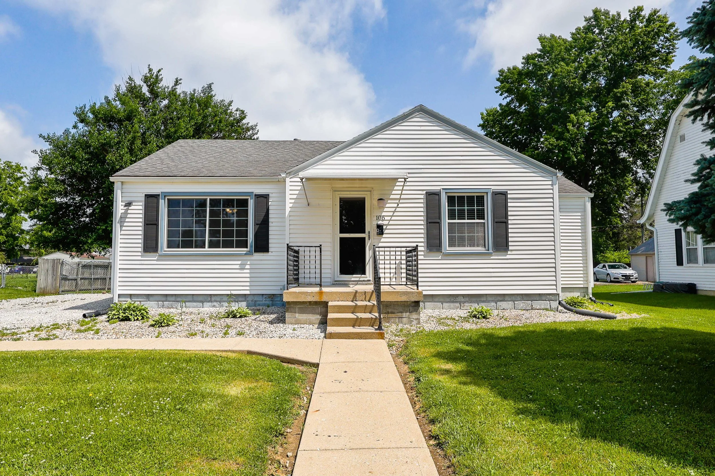 Front view of a small, white, single-story house with a gray roof, black shutters, and a set of steps leading to the front door. There is a green lawn, trees in the background, and a partly cloudy sky.