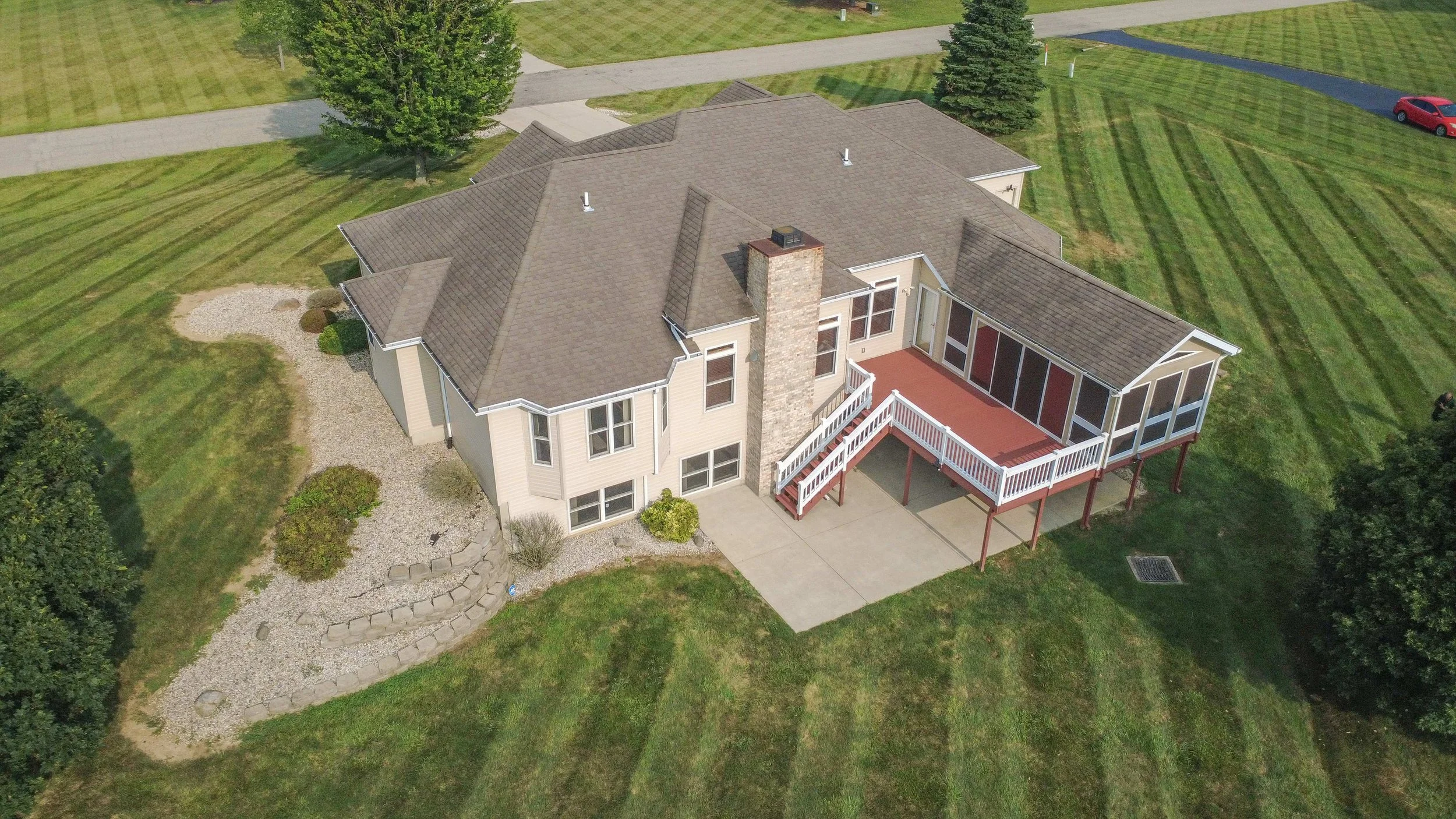 Aerial view of a two-story house with a brown roof, surrounded by a well-maintained lawn with striped grass, trees, and a paved driveway.