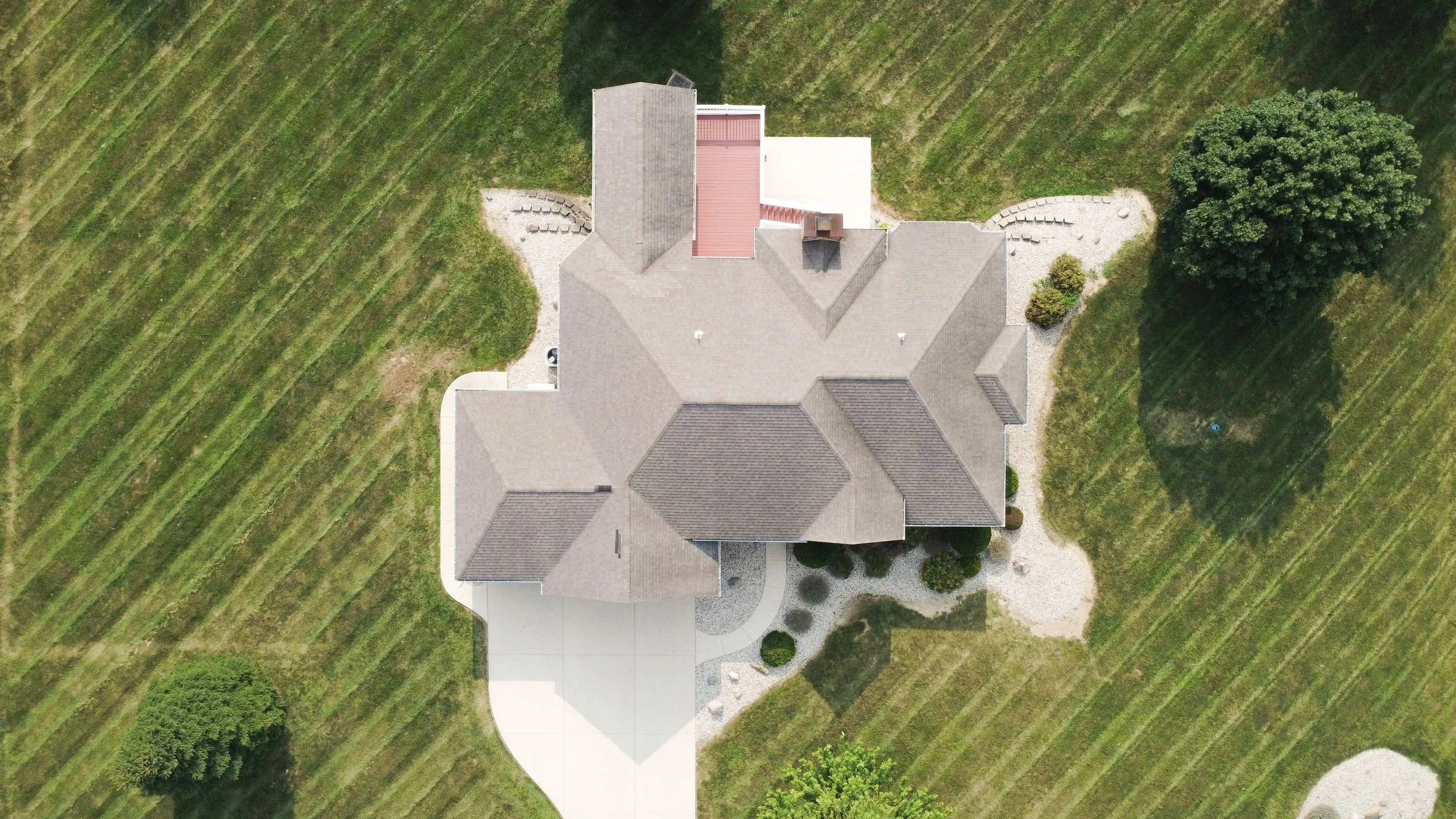 An aerial view of a large house with a gray roof, surrounded by a landscaped yard with trees, bushes, and a gravel pathway, and open green fields.