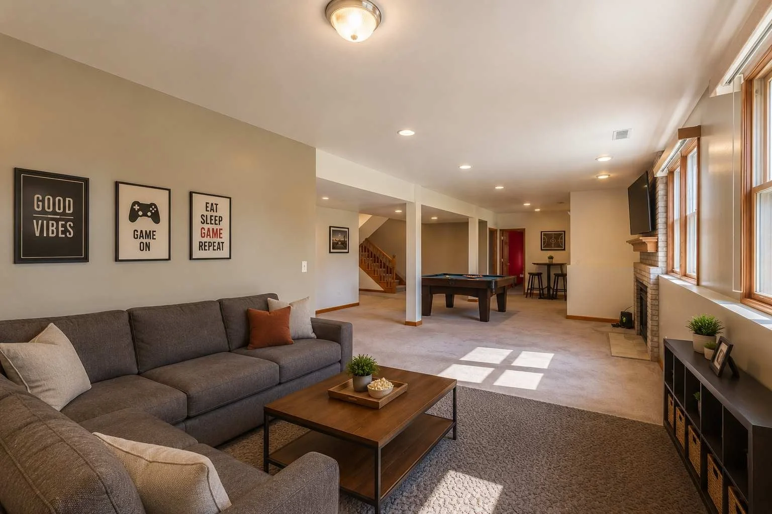 Living room with a gray sofa, wooden coffee table, wall art with gaming and positive messages, fireplace, and large windows, illuminated by natural light.