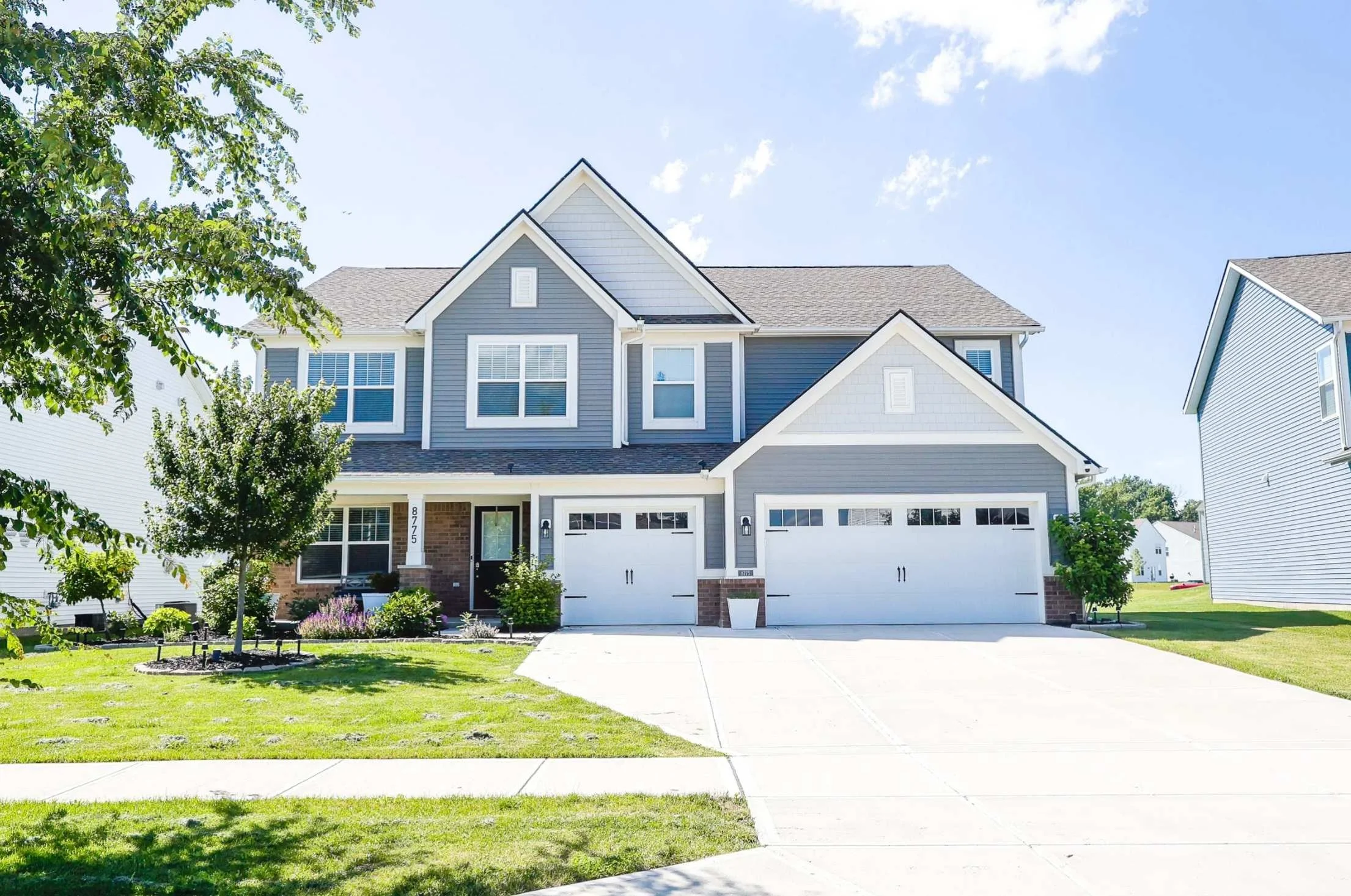 A two-story house with blue siding, white trim, and a gray roof. It has three front-facing windows and a white garage door. The front yard has green grass, small trees, and plants, with a concrete driveway leading to the garage. The sky is clear with