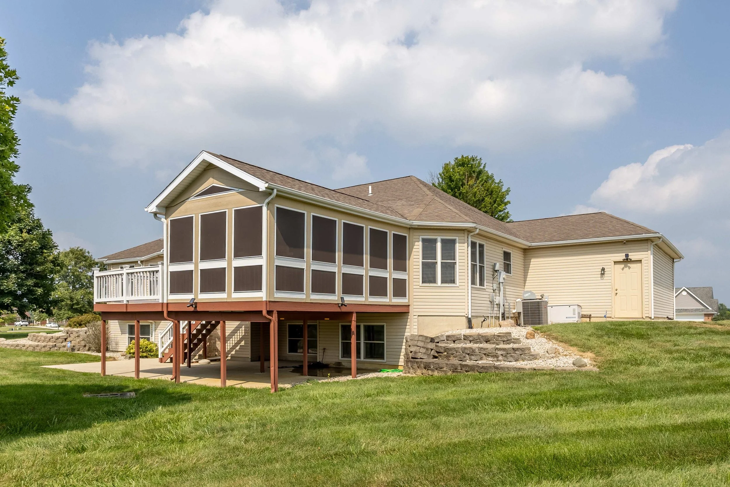 Rear view of a two-story house with beige siding, screened porch, and a brown roof. There is a grassy yard with a stone border, a staircase leading to the porch, and some outdoor units and pipes on the side.