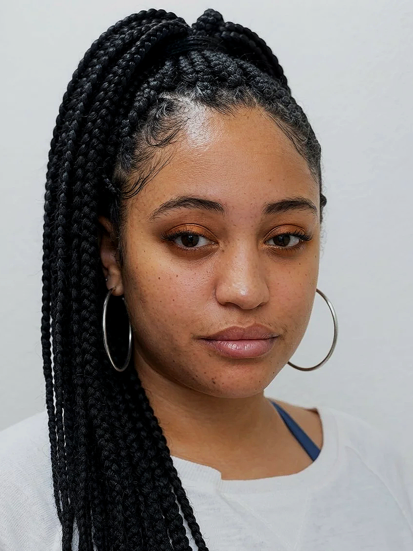 A young woman with braided hairstyle, hoop earrings, and makeup, looking at the camera against a plain white background.
