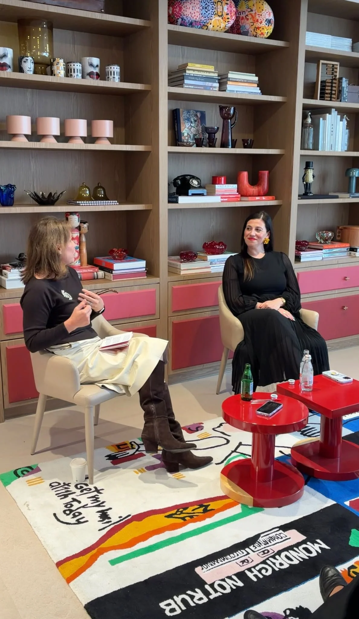 A woman in black dress with yellow earrings sitting in a beige chair, and a girl with a black shirt and beige skirt sitting in a similar chair, engaged in conversation in a modern, colorful living room. Behind them is a wooden bookshelf filled with decorative items, books, and colorful objects. On the floor, there is a vibrant rug with various patterns and text, and red tables with bottles, cups, and a phone.