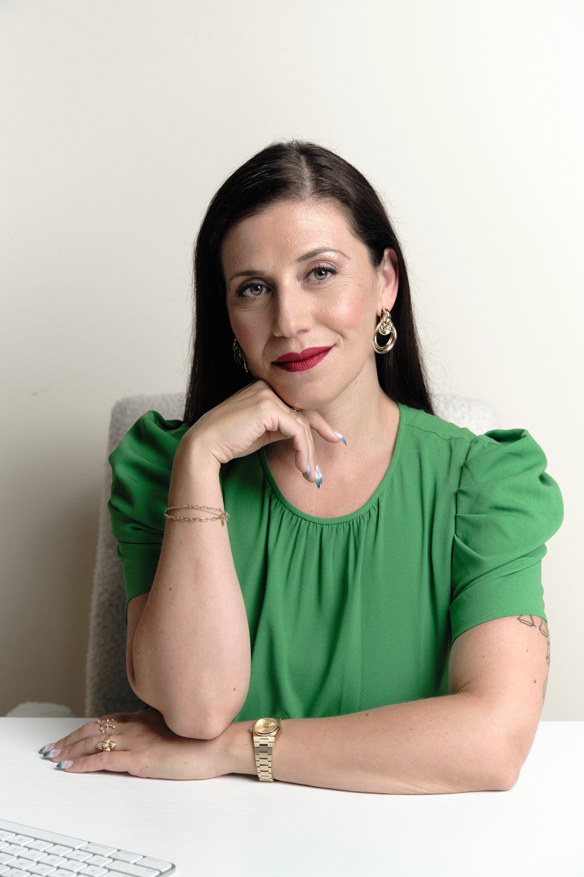 A woman with dark brown hair and light skin wearing a green top, sitting at a white desk, resting her chin on her hand, looking confidently at the camera, with a white background.