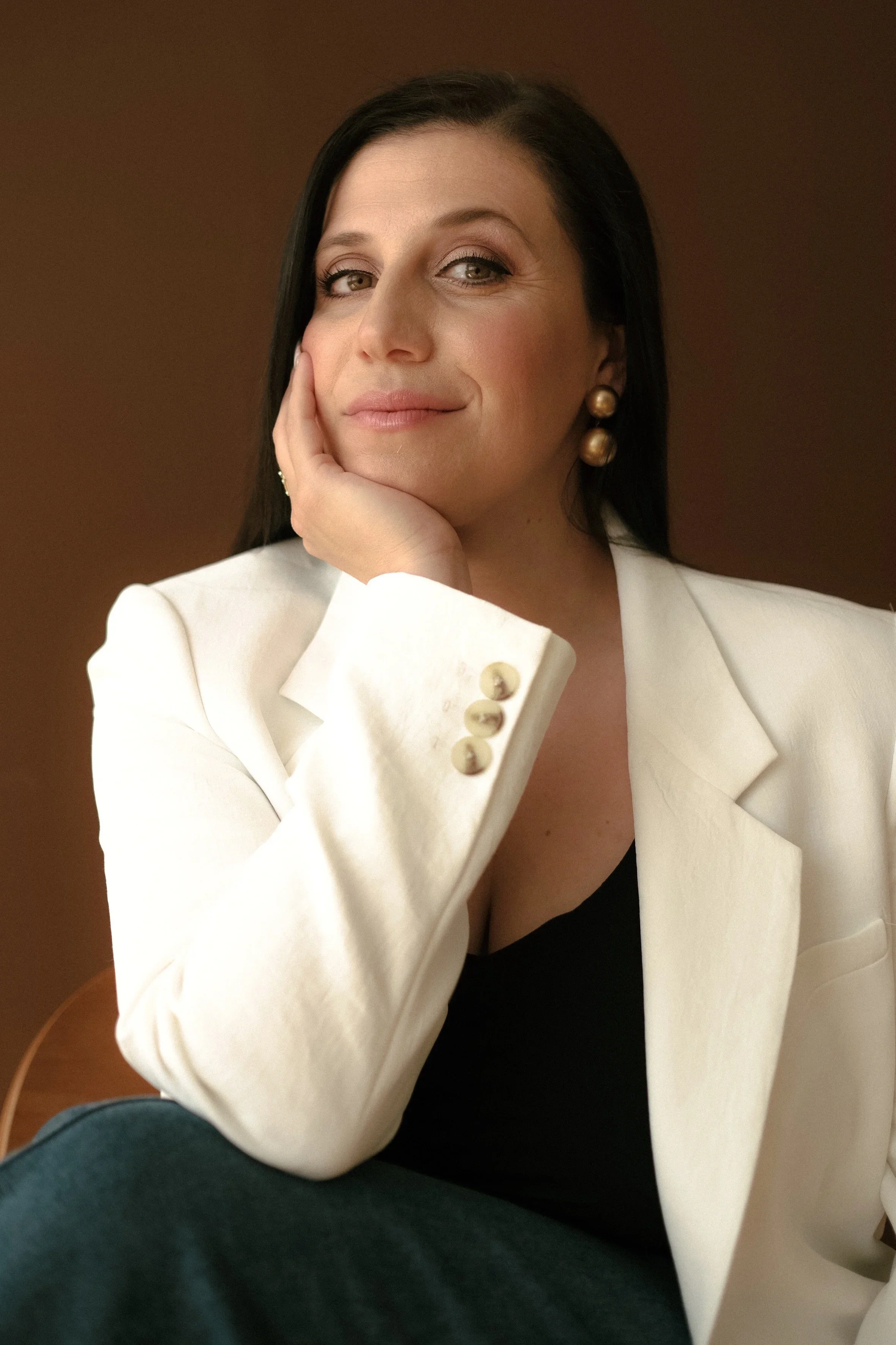 A woman with dark hair and green eyes sitting on a chair, resting her face on her hand, wearing a white blazer with gold buttons, a black top, and gold earrings, against a dark plain background.