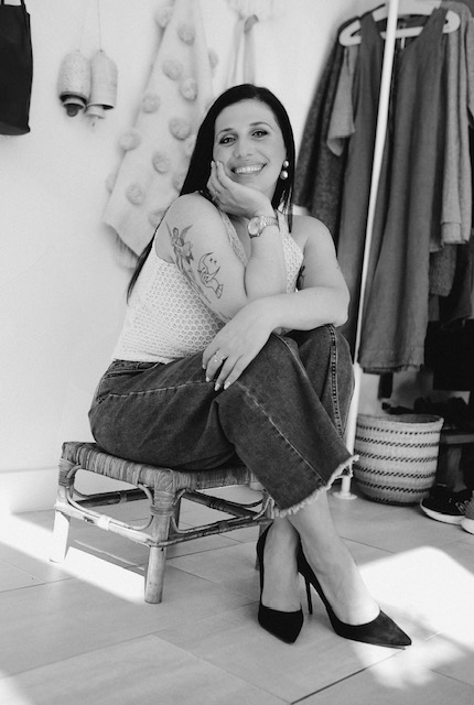 Woman sitting on a chair, smiling, with tattoos on her arm, in a room with hanging clothes and household items in the background.