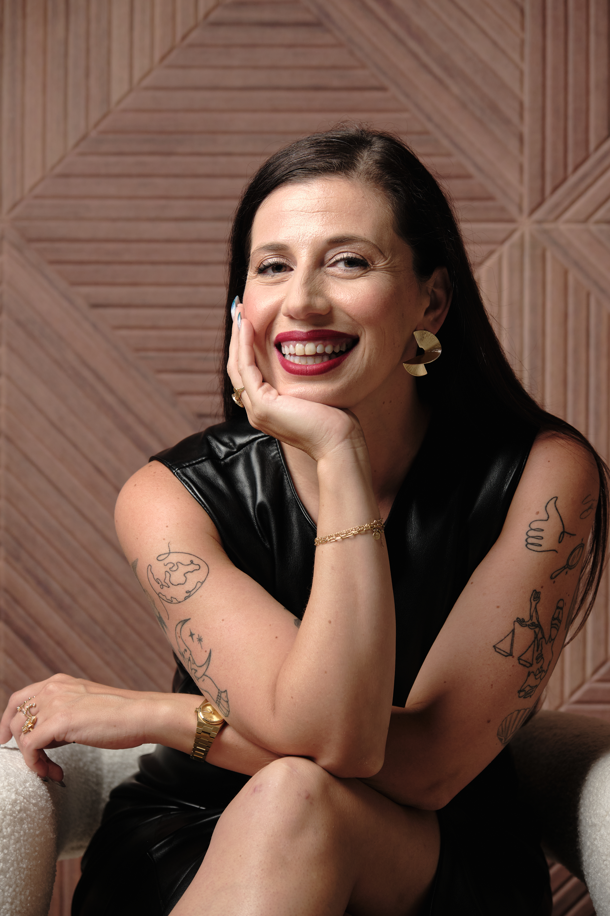 A woman with dark hair, tattoos on her arms, wearing large gold earrings, a gold bracelet, and a leather top, smiling with her hand on her face, sitting on a textured cream-colored chair against a wooden wall background.