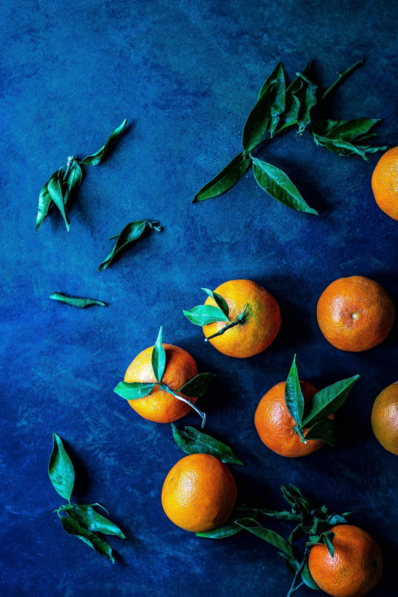 A group of ripe oranges with green leaves and some scattered leaves on a dark blue textured surface.