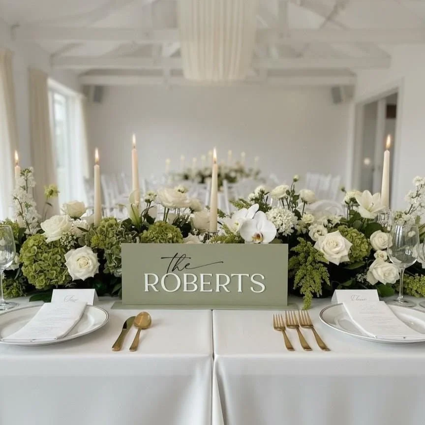 Sage acrylic top table sign with a raised white surname displayed among wedding flowers