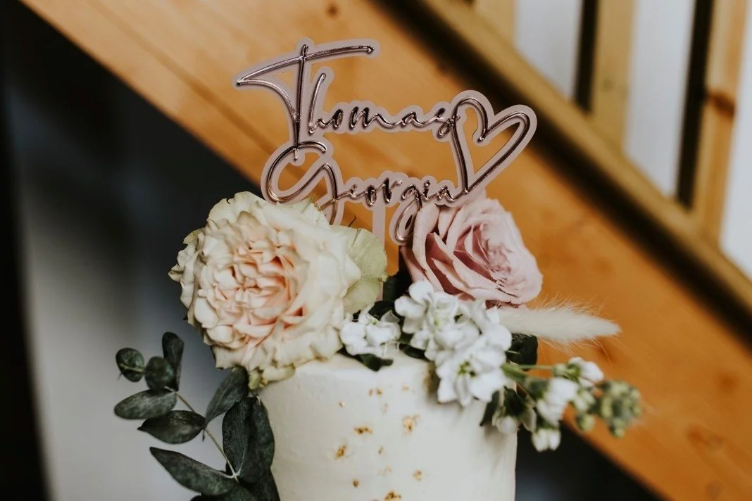 Rose-gold personalised names and heart acrylic topper displayed on a floral wedding cake