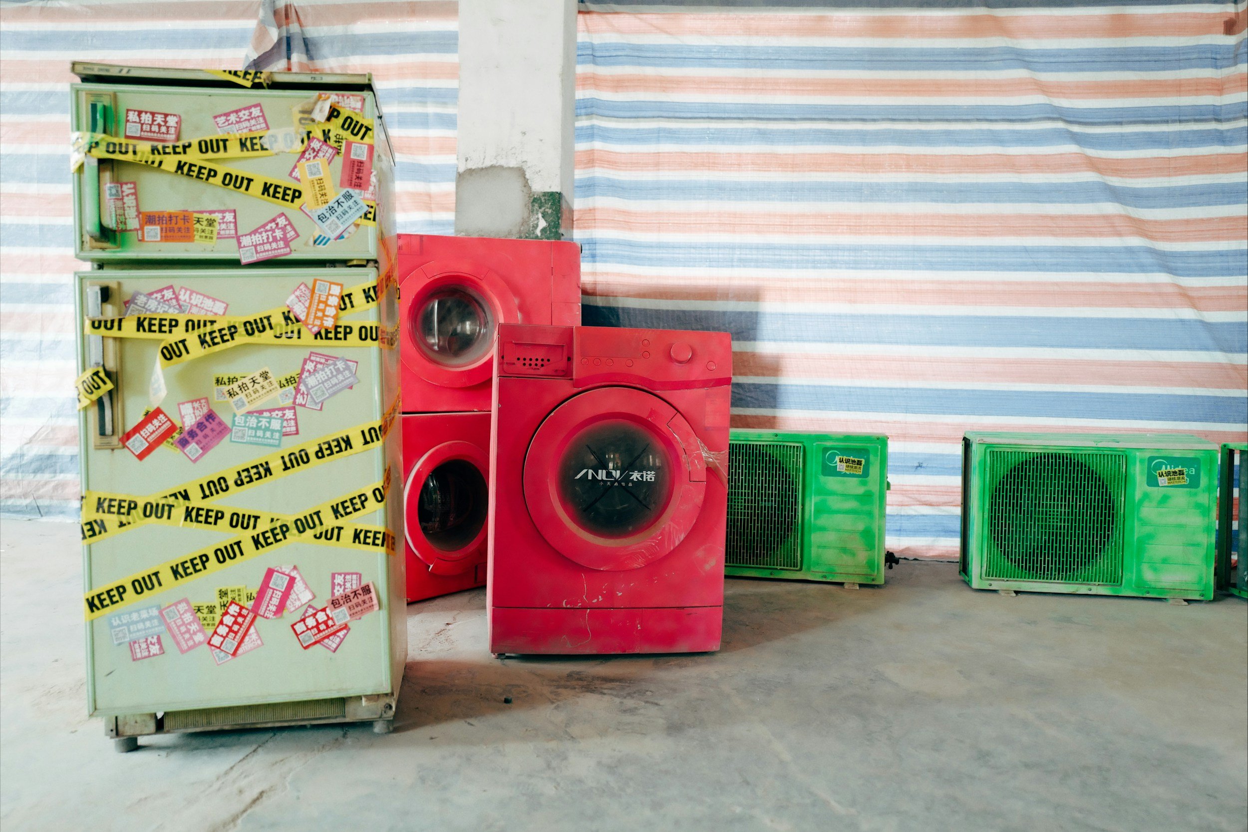 A row of appliances including a green refrigerator covered with 'Keep Out' and other stickers, a red washing machine, a red dryer stacked on top of the washer, and two green air conditioning units set against a striped fabric backdrop.