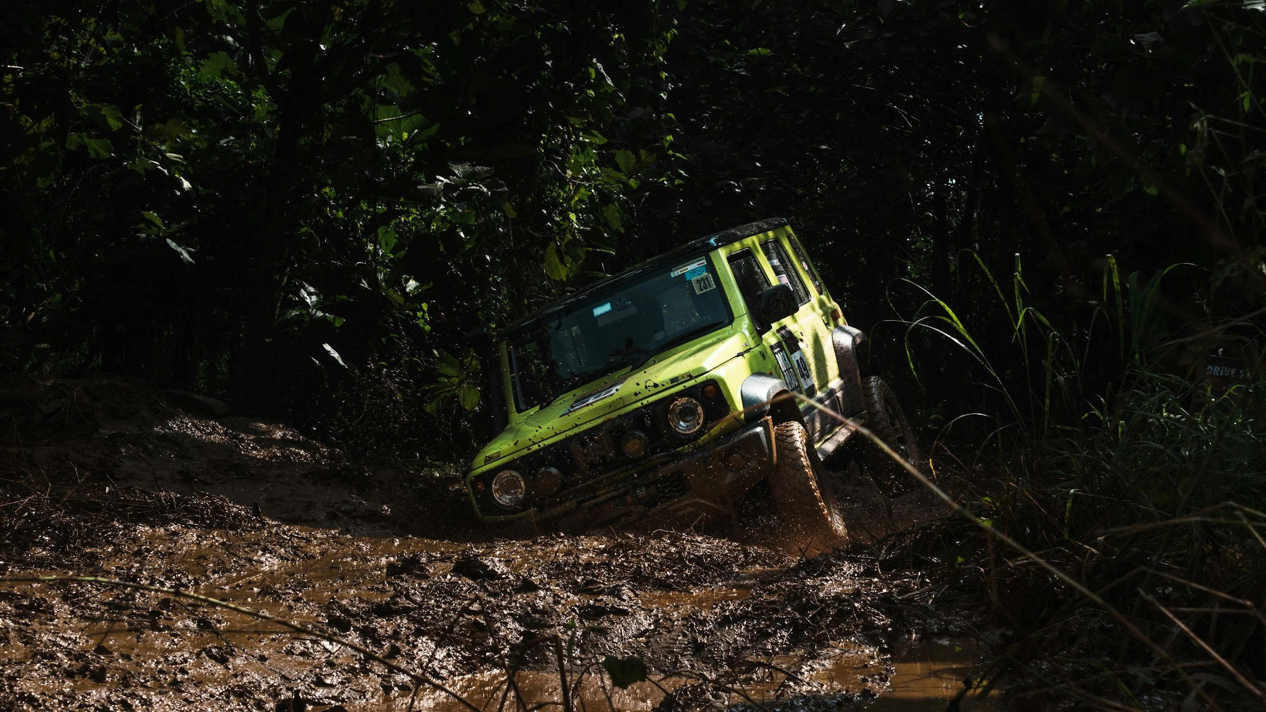A green Jeep vehicle navigating through a muddy, off-road trail surrounded by dense jungle foliage.