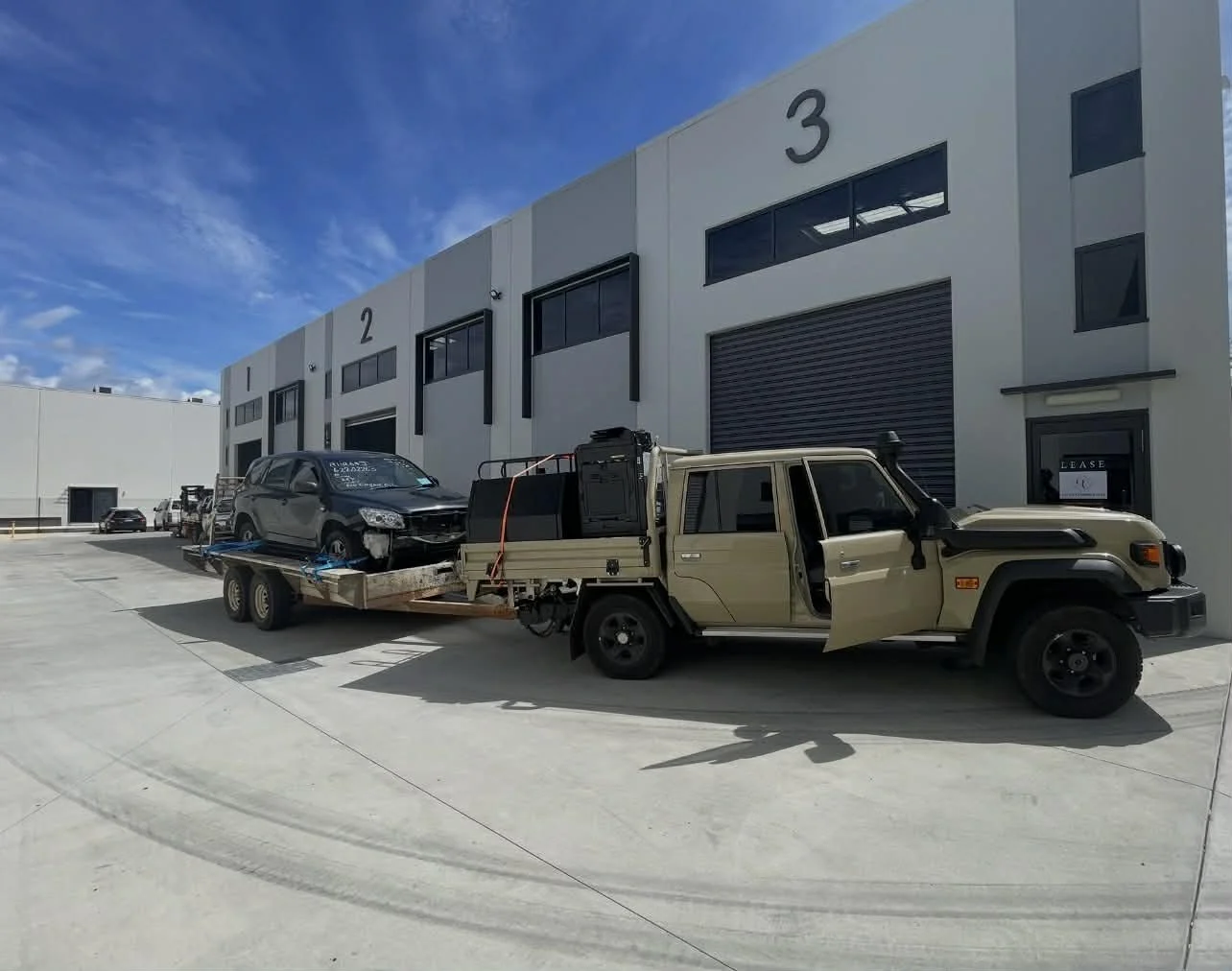 A tow truck parked outside a commercial warehouse with large garage doors labeled 1, 2, and 3, carrying a damaged gray SUV.