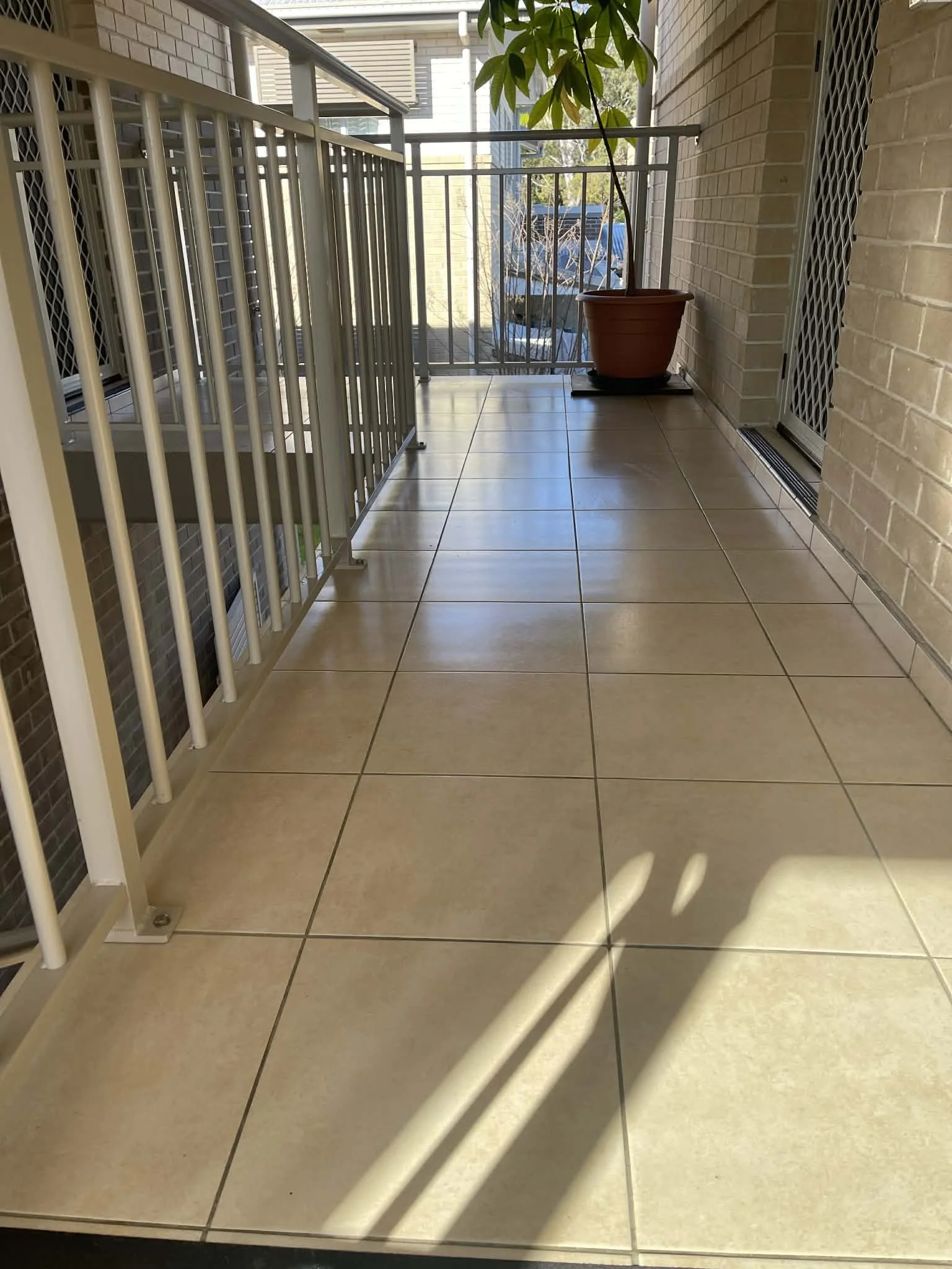 A balcony with beige tiled flooring, a white metal railing on the left side, and a brown planters with a plant on the right side against a brick wall. Shadows from the balcony railing are cast on the floor, and there is a sliding glass door on the right side.