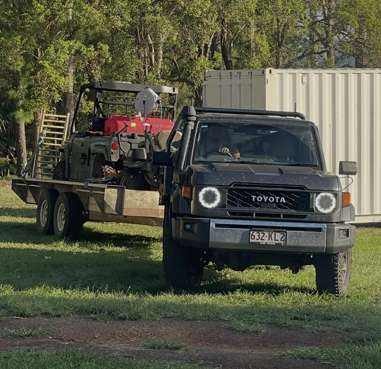 A black Toyota pickup truck parked on grass, carrying a golf cart, a red fuel container, and other equipment, with trees and a white container in the background.
