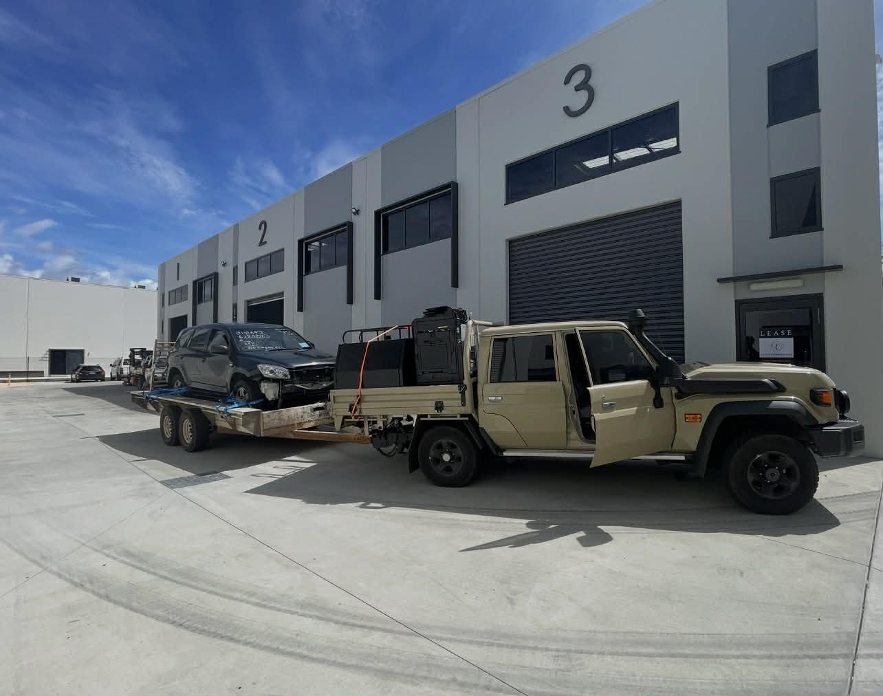 A tow truck carrying a black SUV with front damage in front of a commercial industrial building with numbered bays.