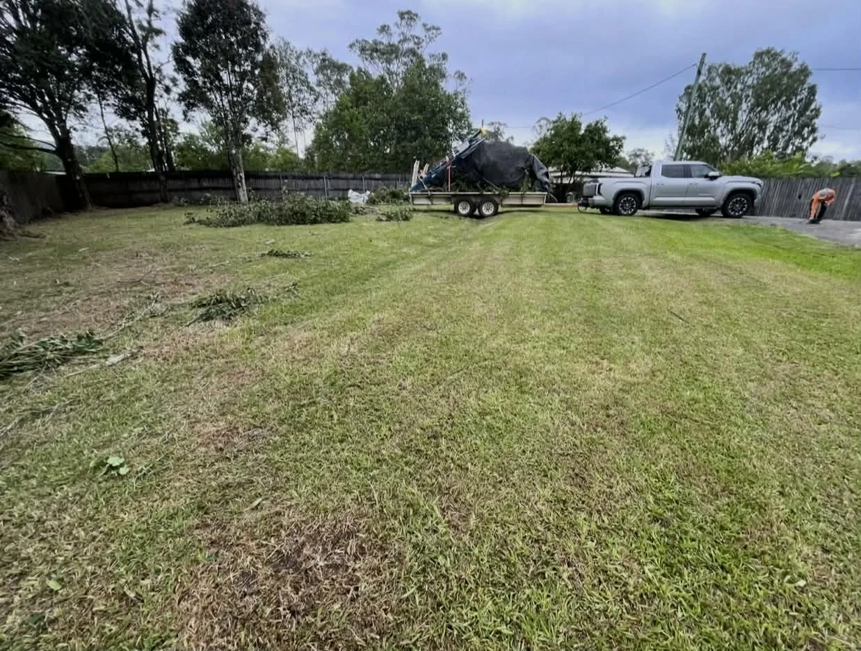 A backyard with grass, trees, a wooden fence, a trailer with debris, a gray pickup truck, and a person working near the fence.