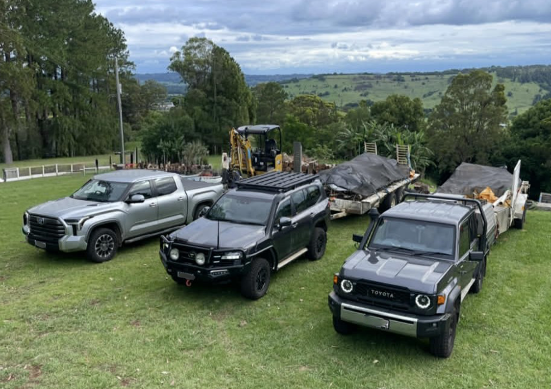 Three pickup trucks and a small excavator parked on a grassy lawn with trees and a rural landscape in the background.