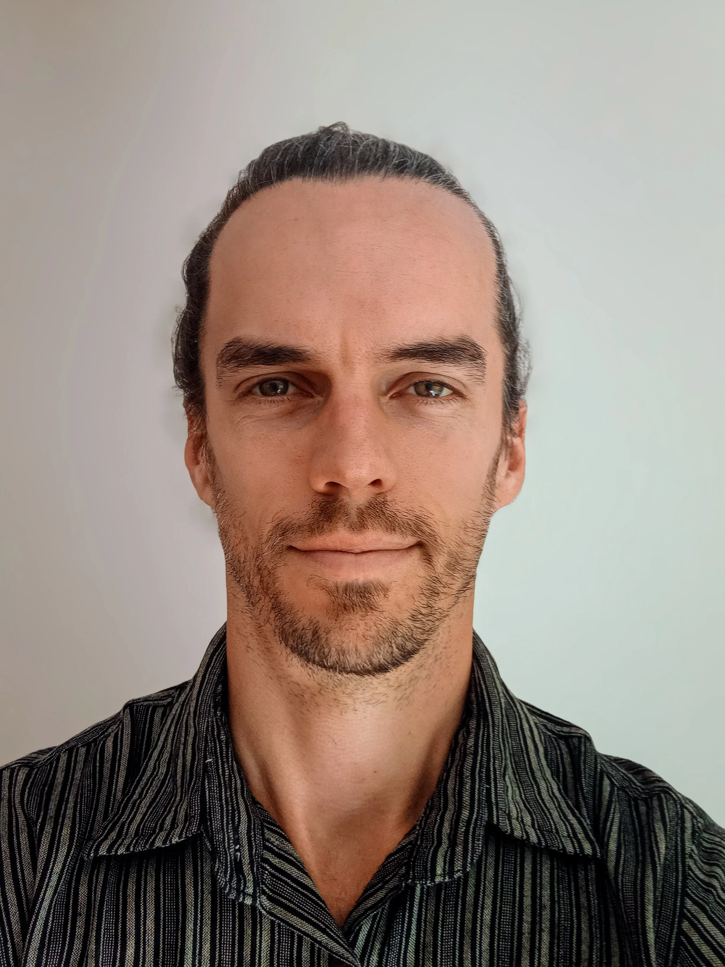 Close-up of a man with dark hair, blue eyes, and facial hair, wearing a black and gray striped shirt, standing in front of a plain light-colored wall.