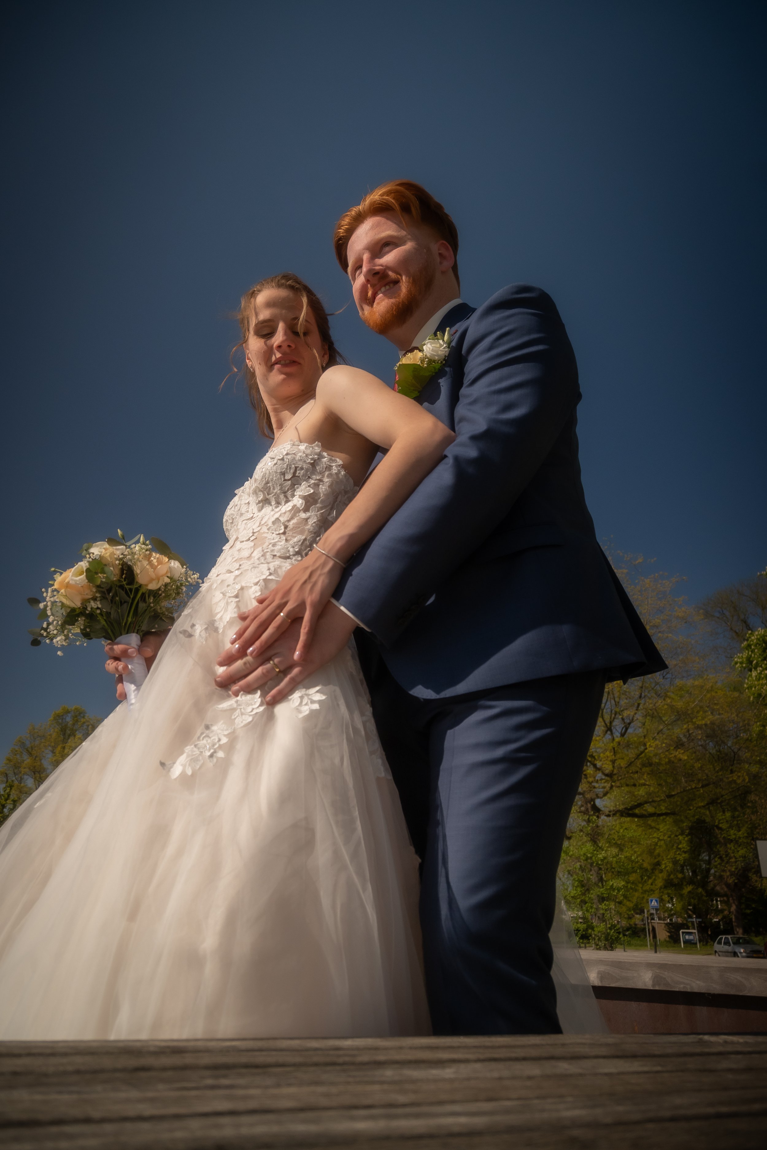Een bruid en bruidegom die samen poseren buiten op een dag met een heldere blauwe lucht, de bruid vasthoudend een boeket bloemen en de bruidegom in een donkerblauw pak.