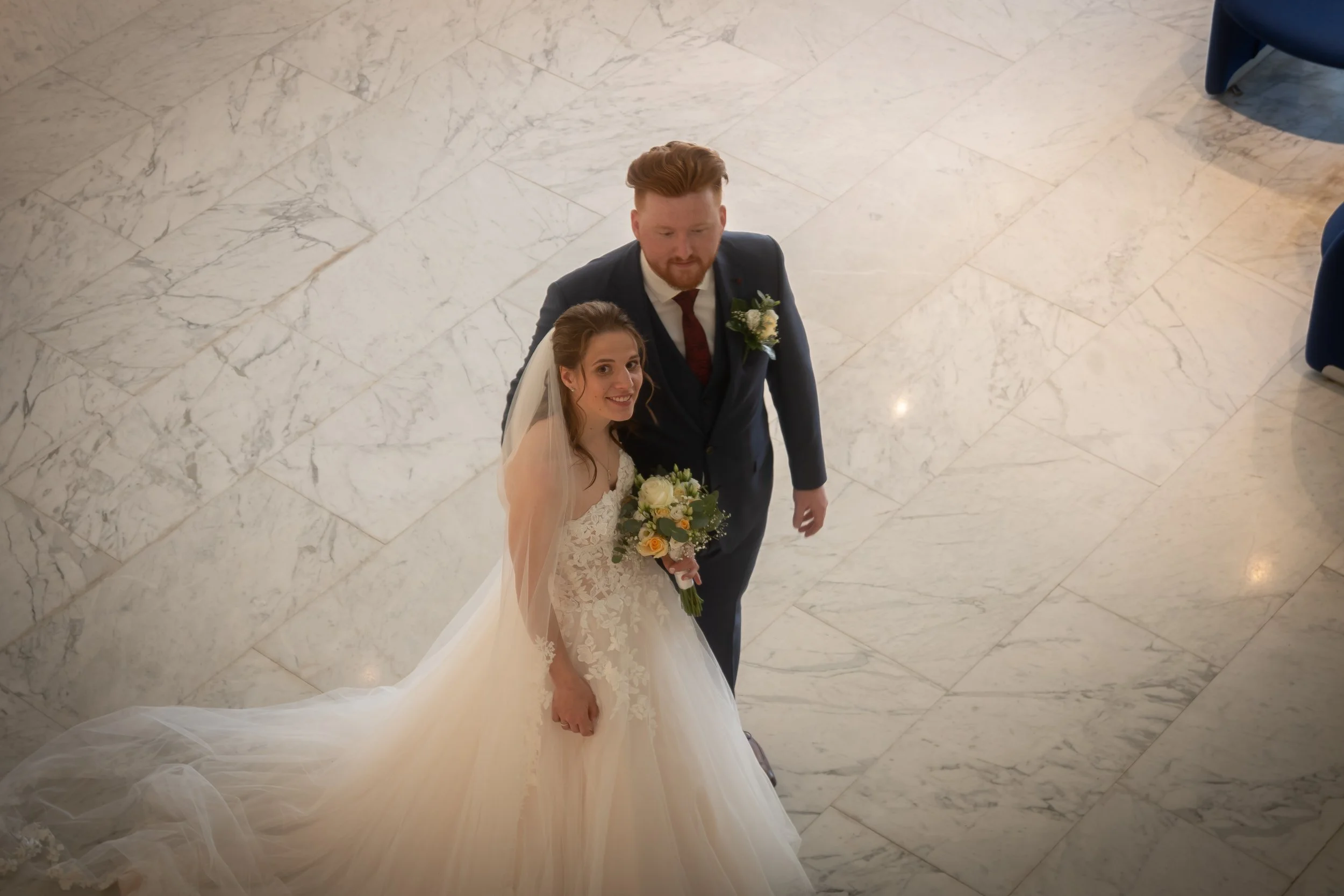 Een pasgetrouwd stel kijkt omhoog naar de camera, de bruid houdt een boeket bloemen vast en draagt een witte jurk met kant, de bruidegom draagt een donker pak met een bloemcorsage, ze staan op een marmeren vloer.