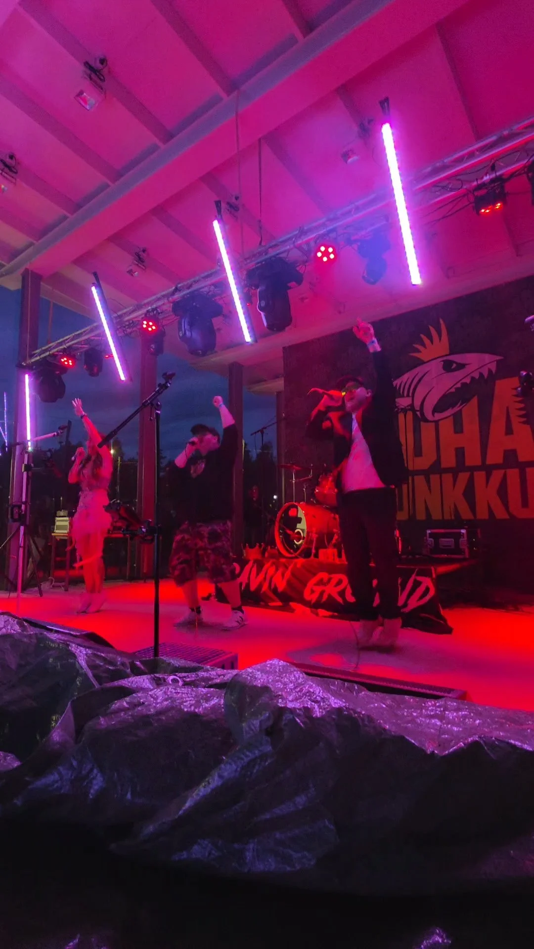 Yleisö nauttii musiikkiesityksestä lavalla, jossa on lavasteet ja valoja, sekä rock-musiikkiaiheinen banneri taustalla.