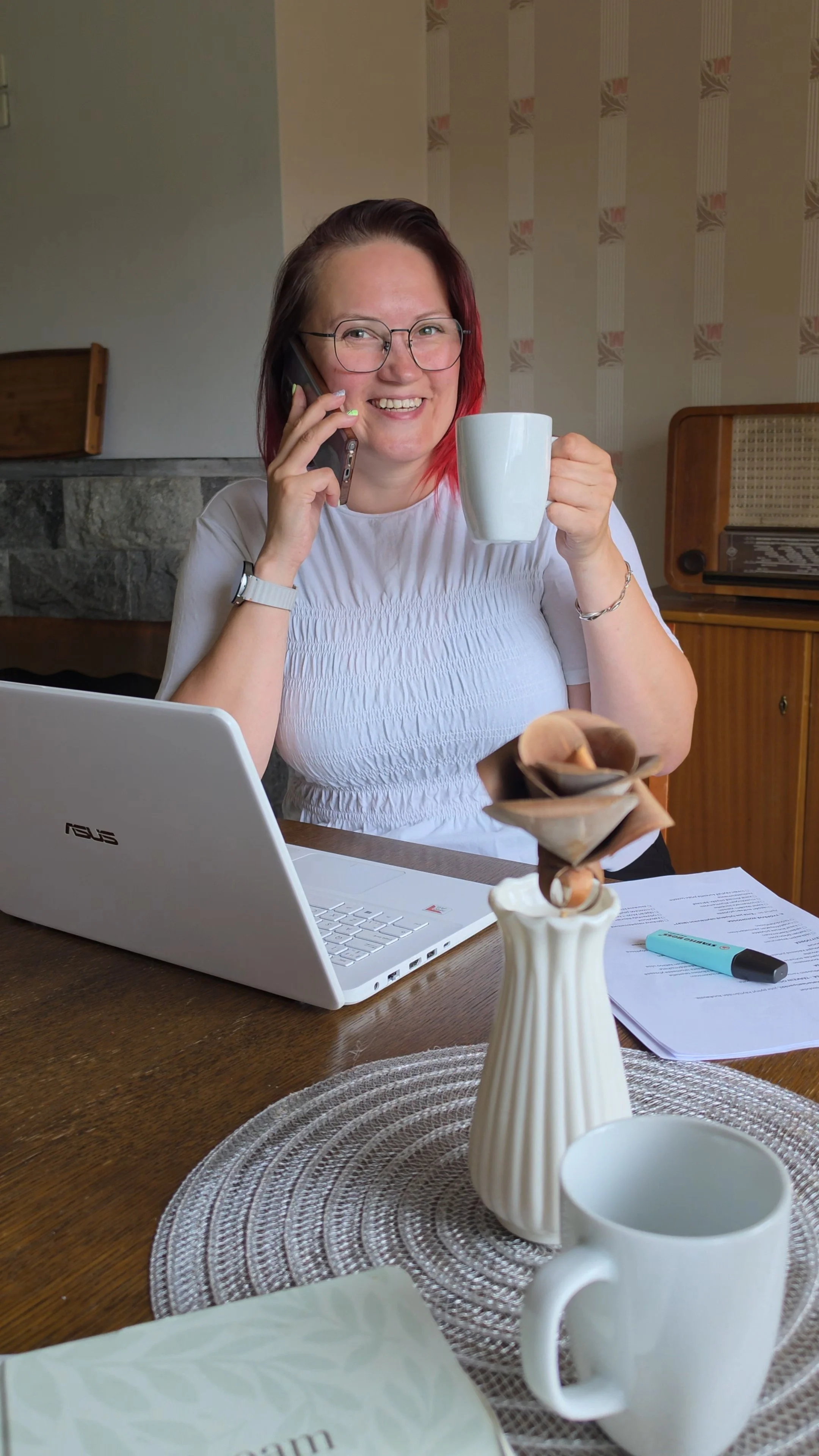 Hymyilevä Hanne, jolla on punaiset hiukset, istuu pöydän ääressä kahvikuppi kädessään ja puhuu puhelimeen, lähellä tietokonetta ja pöytätarvikkeita.