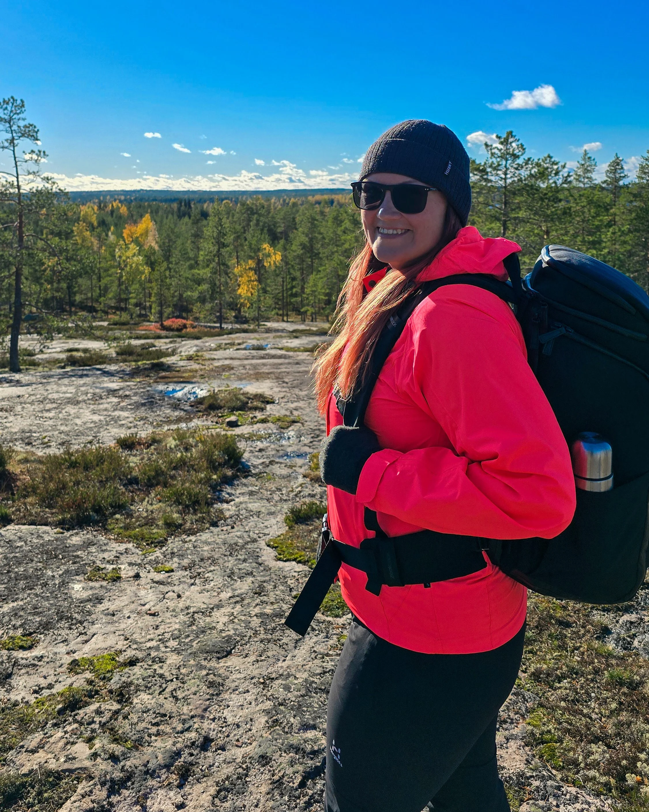 Hanne maastossa urheiluvaatteissa, pipo ja aurinkolasit, on luonnossa metsän ja sinisen taivaan keskellä, nauttii ulkoilmasta.