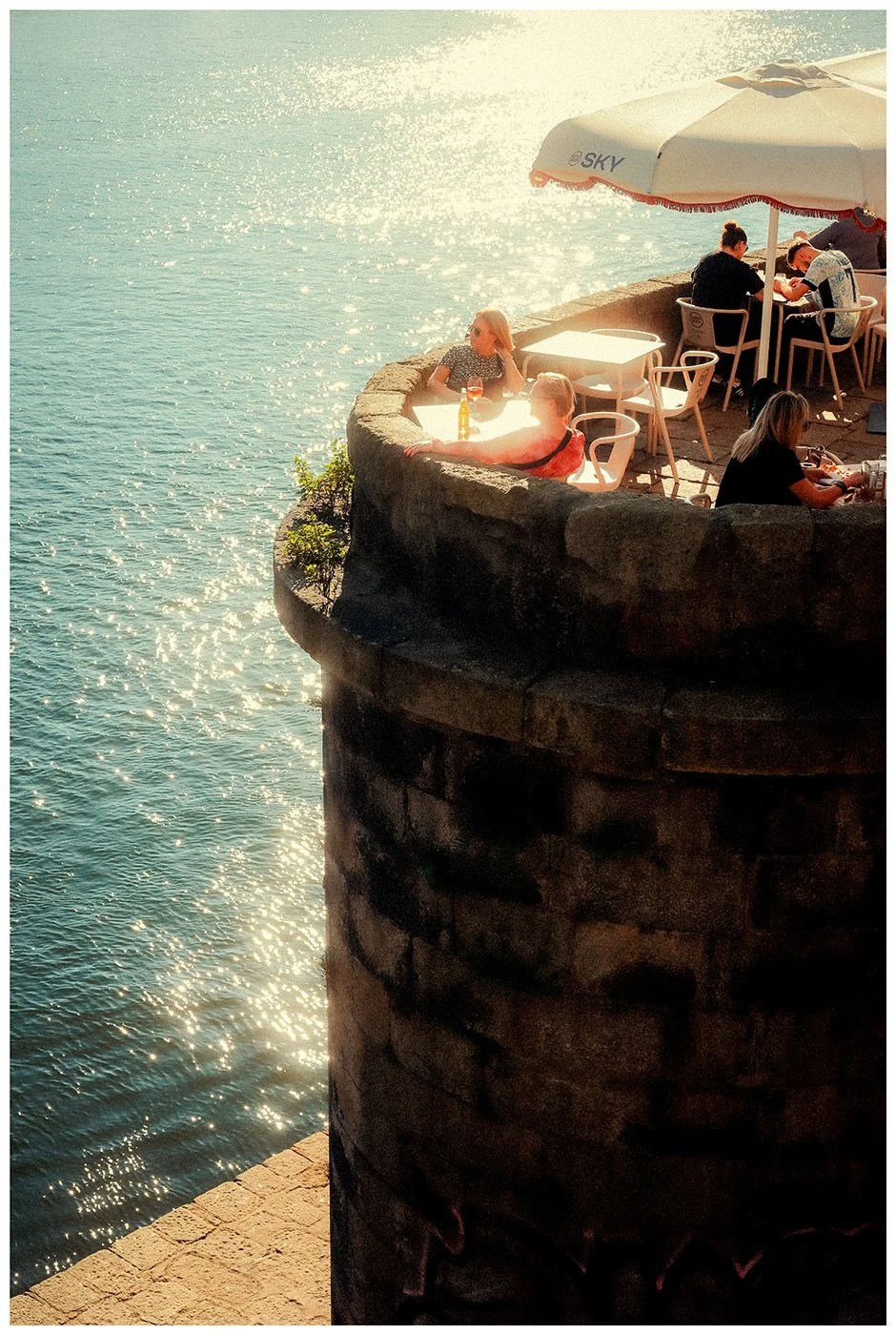People seated at a sunlit café terrace above the Douro River beside an old stone wall in Porto, Portugal.