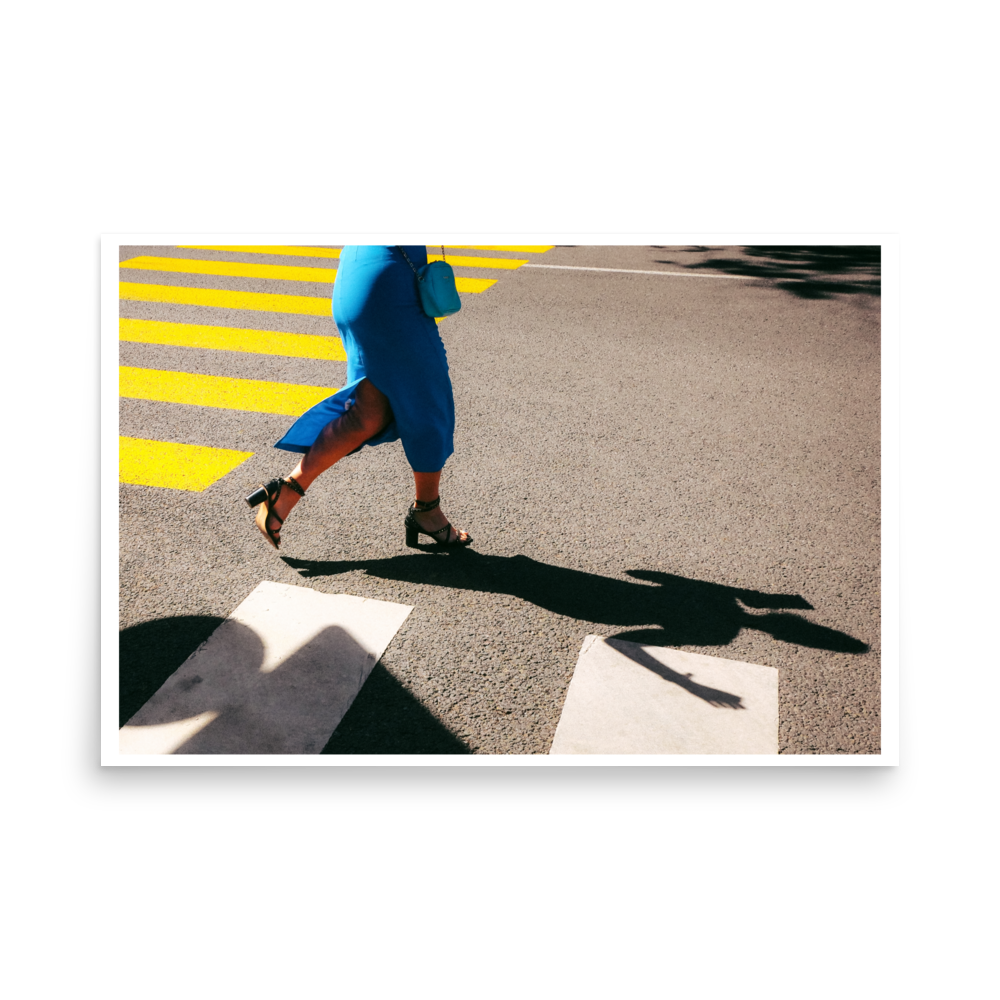 Woman in a blue dress crossing a street with yellow road markings and a long dramatic shadow in bright sunlight.