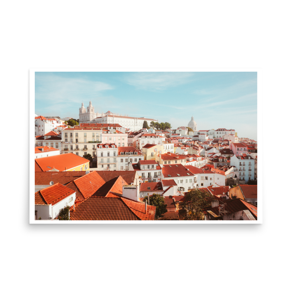 Panoramic view across Lisbon with layered terracotta rooftops, pale buildings and historic church towers beneath a blue sky.