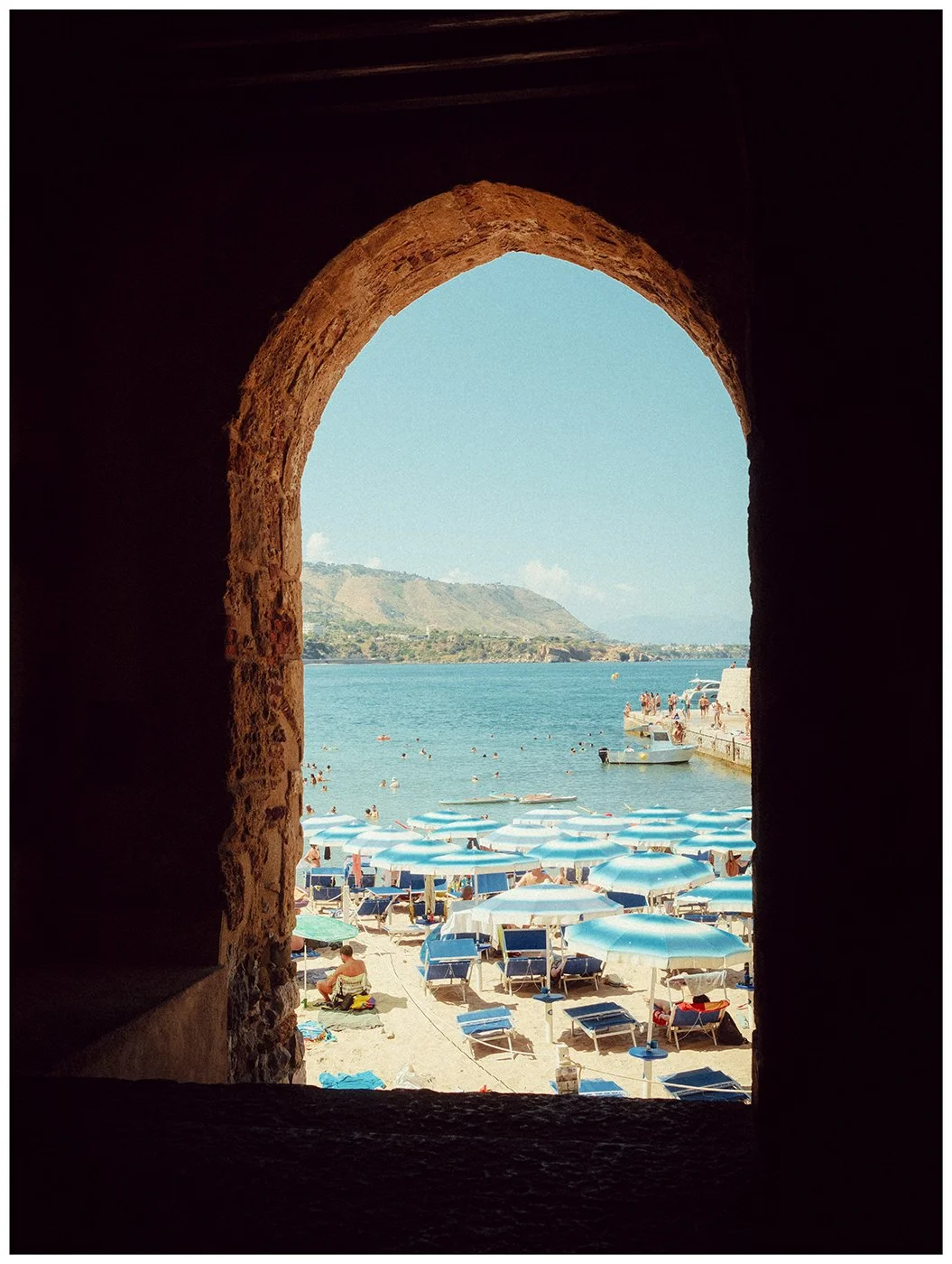 View through a stone arch toward a beach with blue umbrellas and the Mediterranean Sea in Cefalù, Sicily.