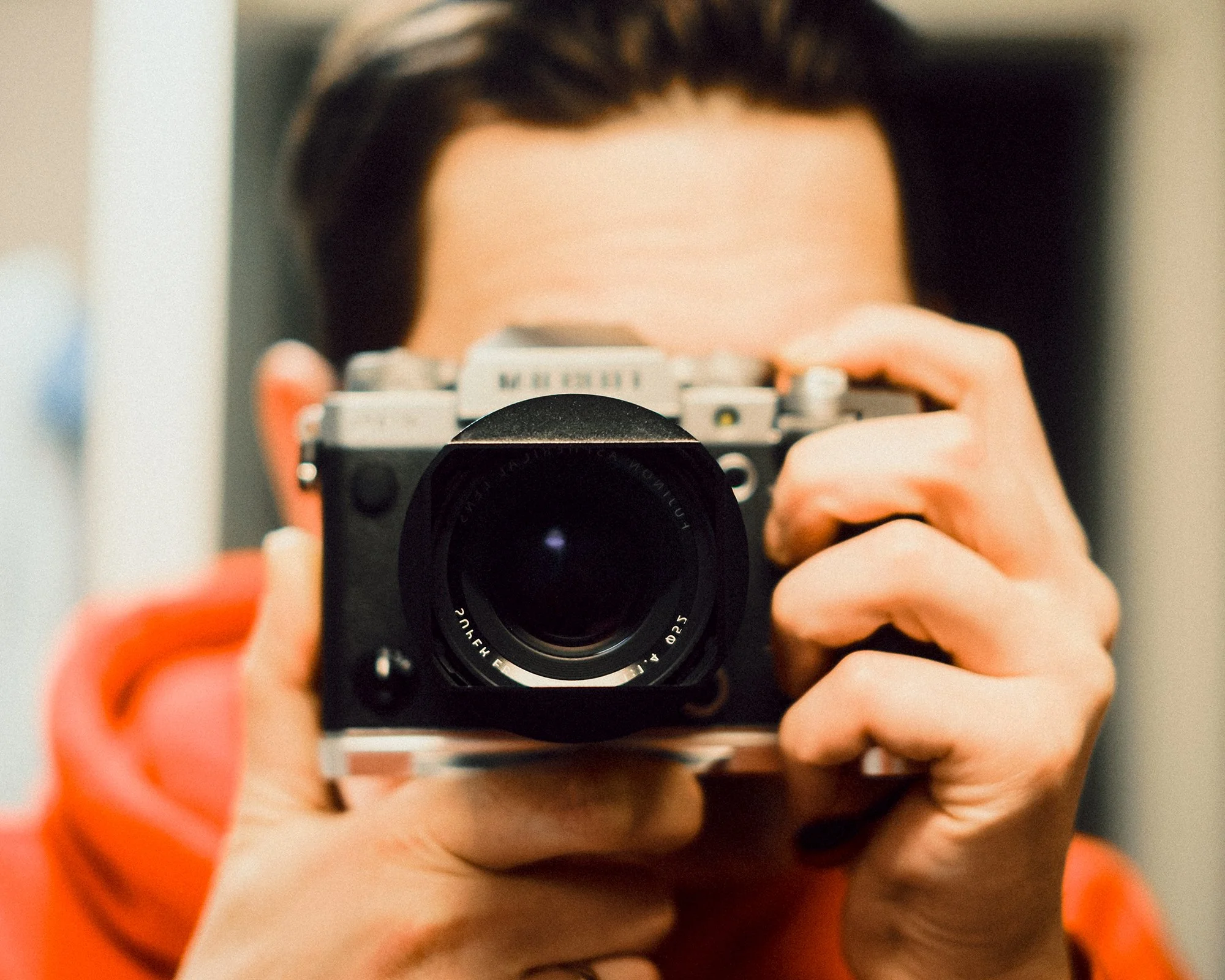 Person holding a camera to their face, wearing an orange top.