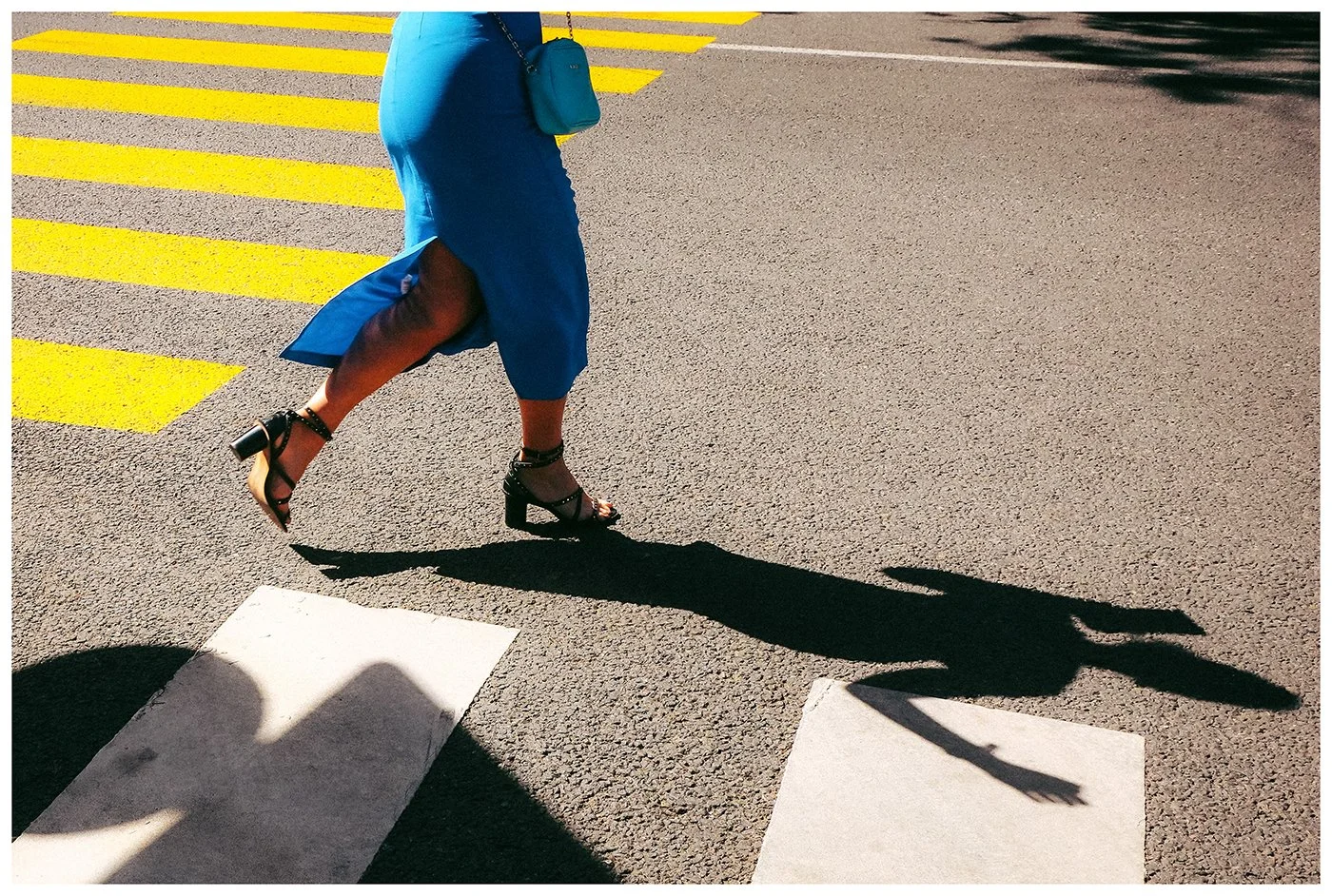 A fleeting street moment where a woman in vivid blue dress crosses bold yellow markings, while her elongated shadow seems to run ahead of her.