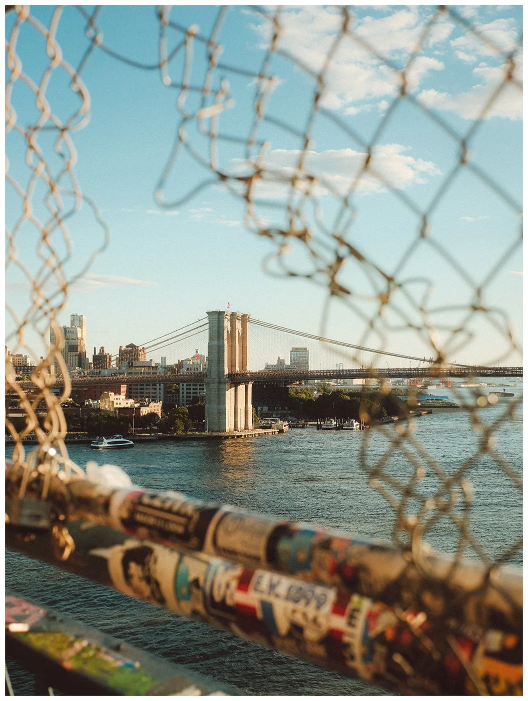 A layered view through the weathered fencing of Manhattan Bridge, framing Brooklyn Bridge beyond.