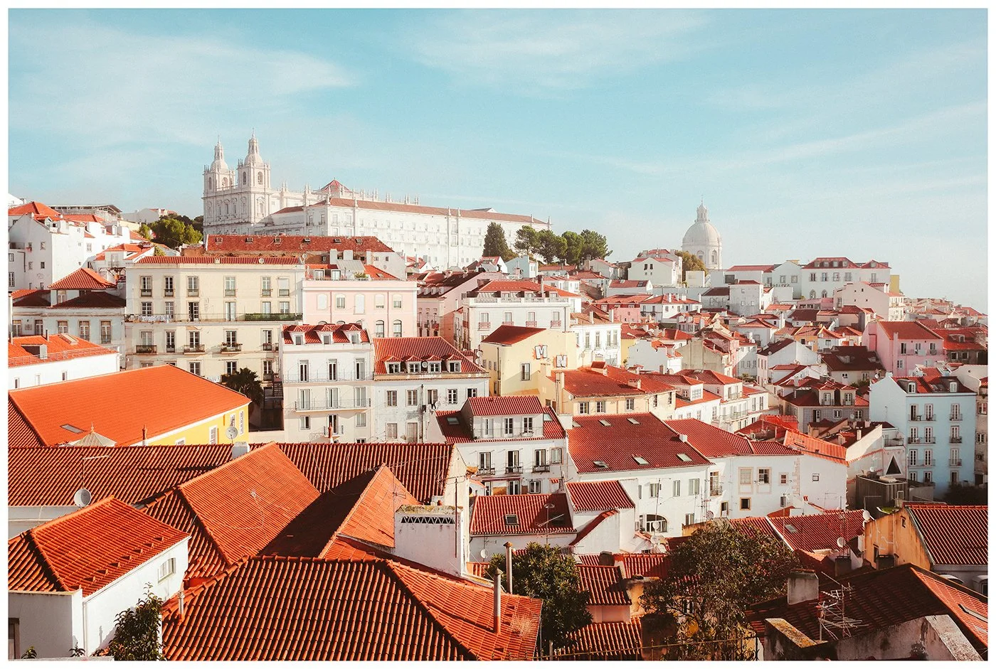 Lisbon unfolds as a layered sea of terracotta rooftops, pale facades and church towers beneath a soft blue sky.